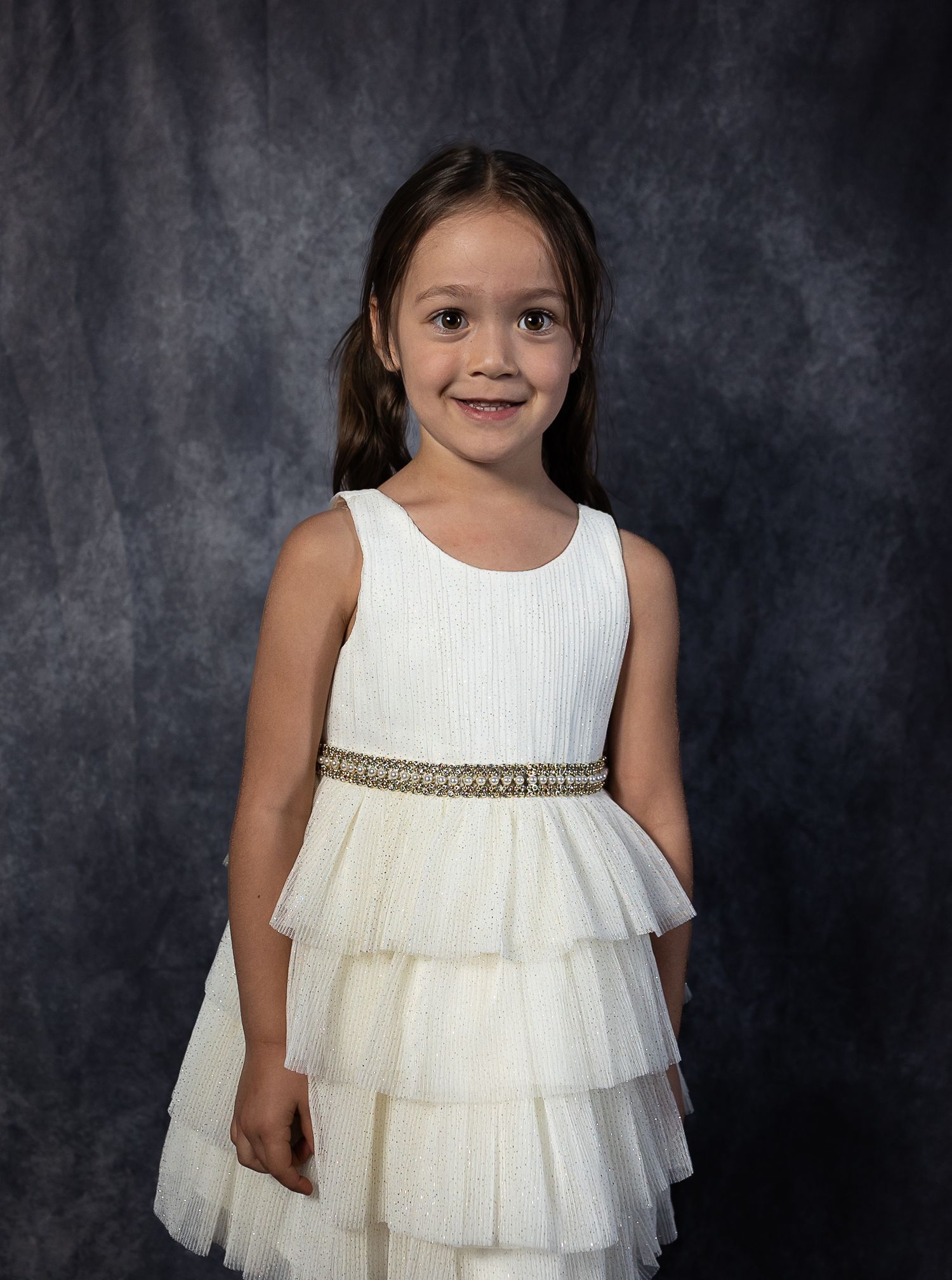 Young girl smiling in a white tiered dress with gold belt, posing for a portrait photo against a gray backdrop.