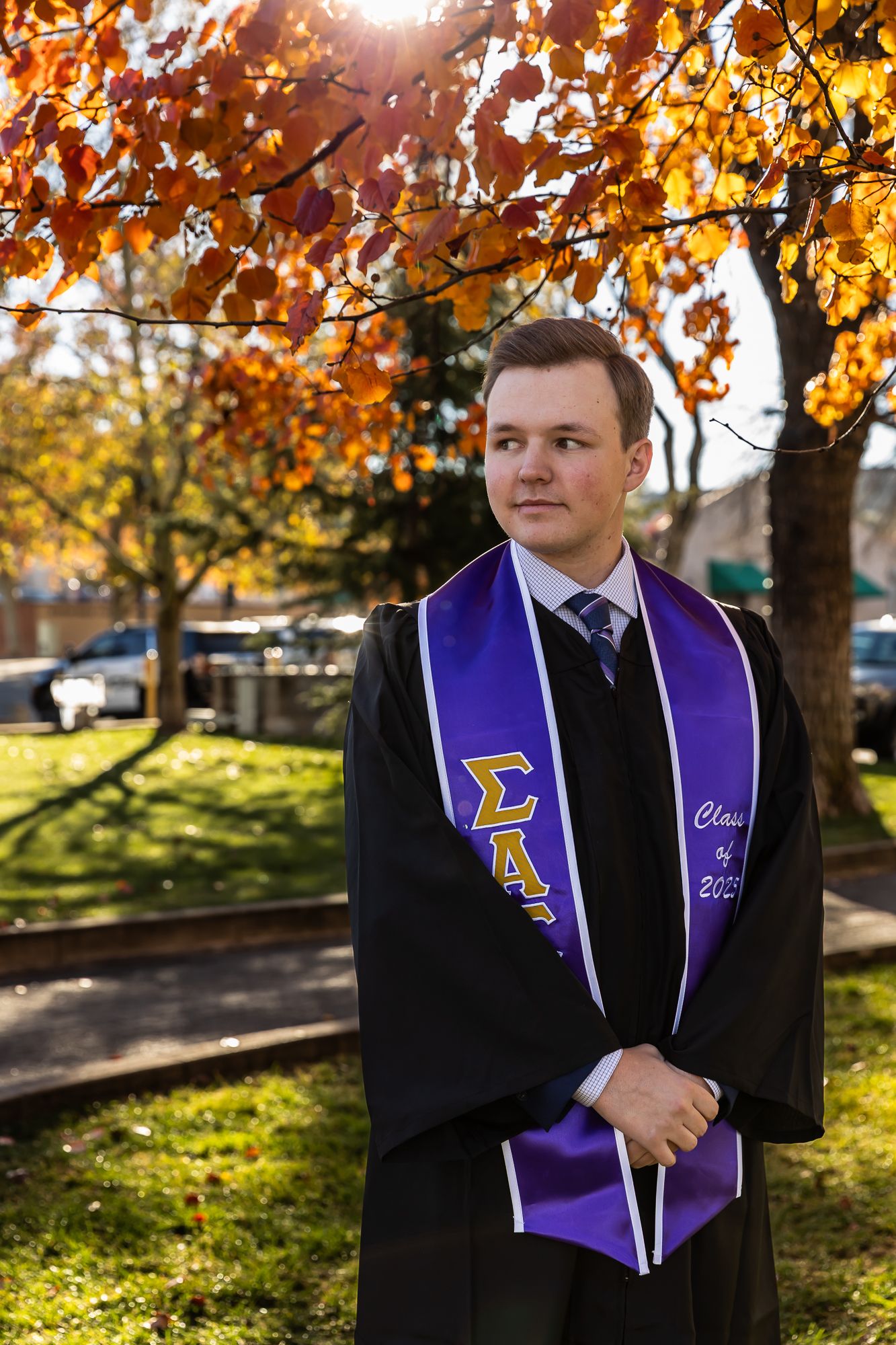 A person wearing a black graduation gown with purple stole stands among autumn trees with golden leaves. arizona Prescott courtyard