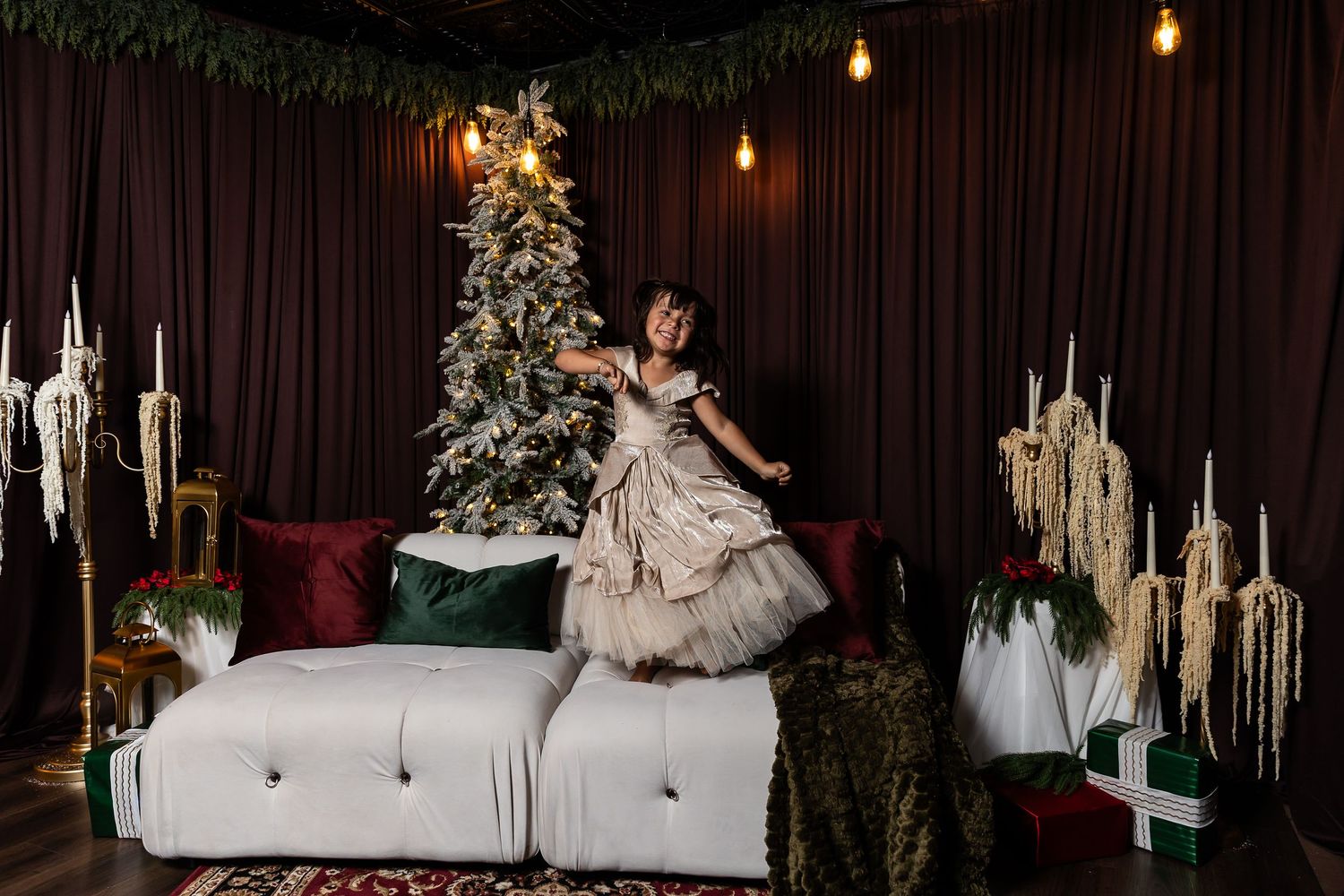 Girl in a tulle dress jumps joyfully on a white tufted sofa beside a decorated Christmas tree in a cozy holiday studio set.