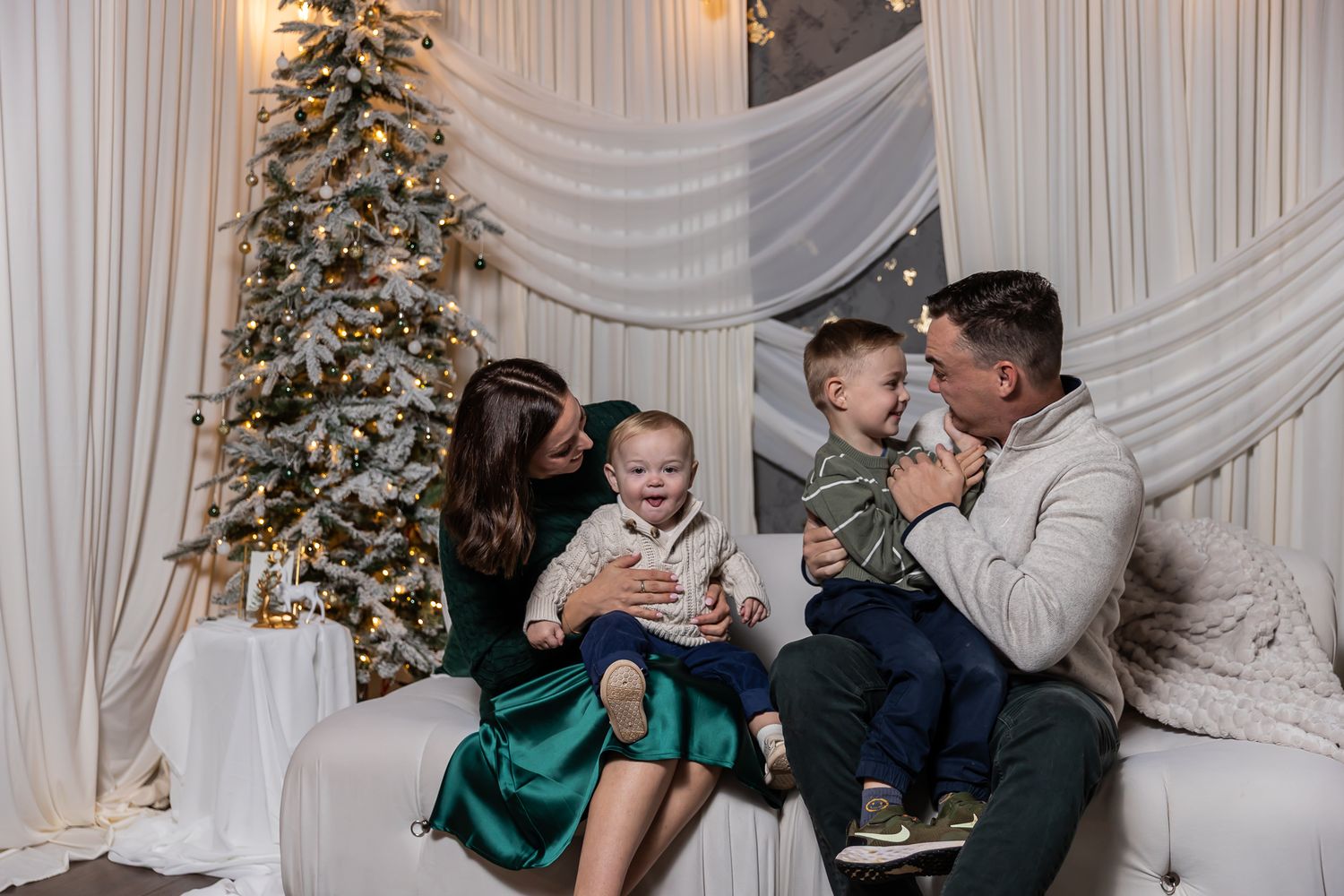 A couple shares a romantic kiss in front of white drapes and decorated Christmas trees at a winter wedding celebration.