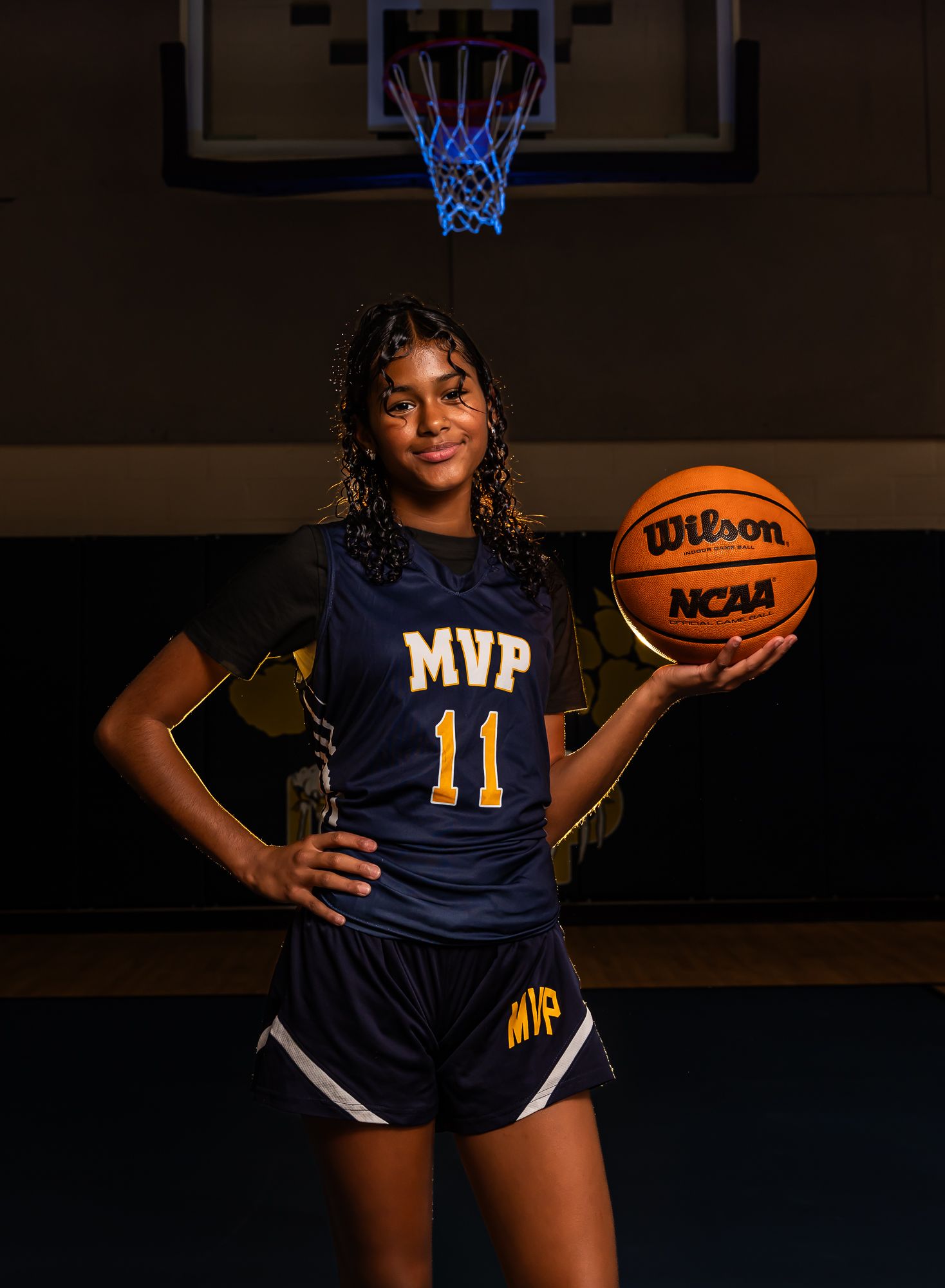 Basketball player in navy MVP jersey poses with NCAA basketball in dark indoor court setting.