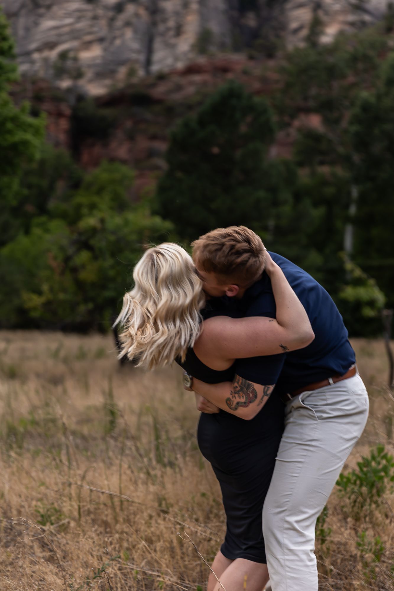 A couple in black and white outfits embrace intimately in a field with rocky cliffs and trees in the background. Sedona Arizona photography