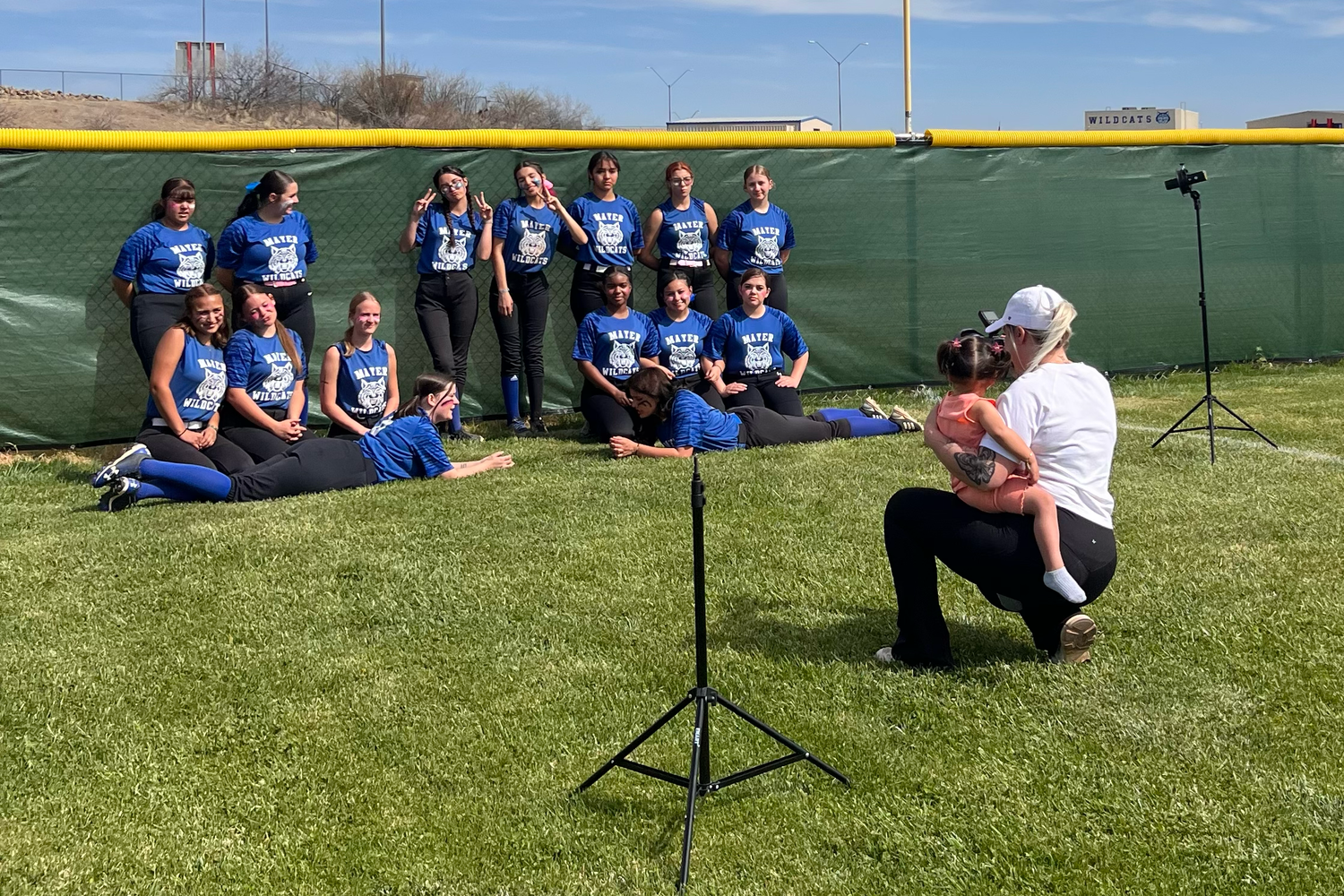 arizona softball players in blue uniforms sit on grass while being photographed during a team photo session on a sunny day.