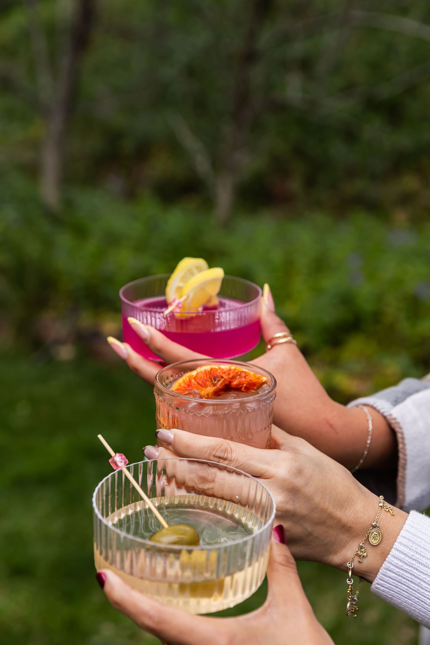 Three colorful cocktails garnished with citrus and olives held by hands with bracelets outdoors.