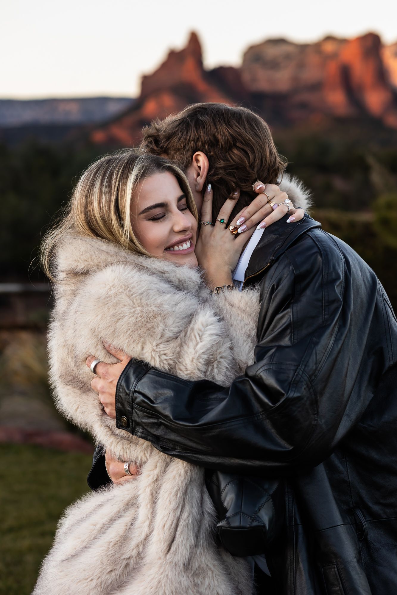 Romantic couple embracing outdoors with person wearing white fur coat against desert landscape.