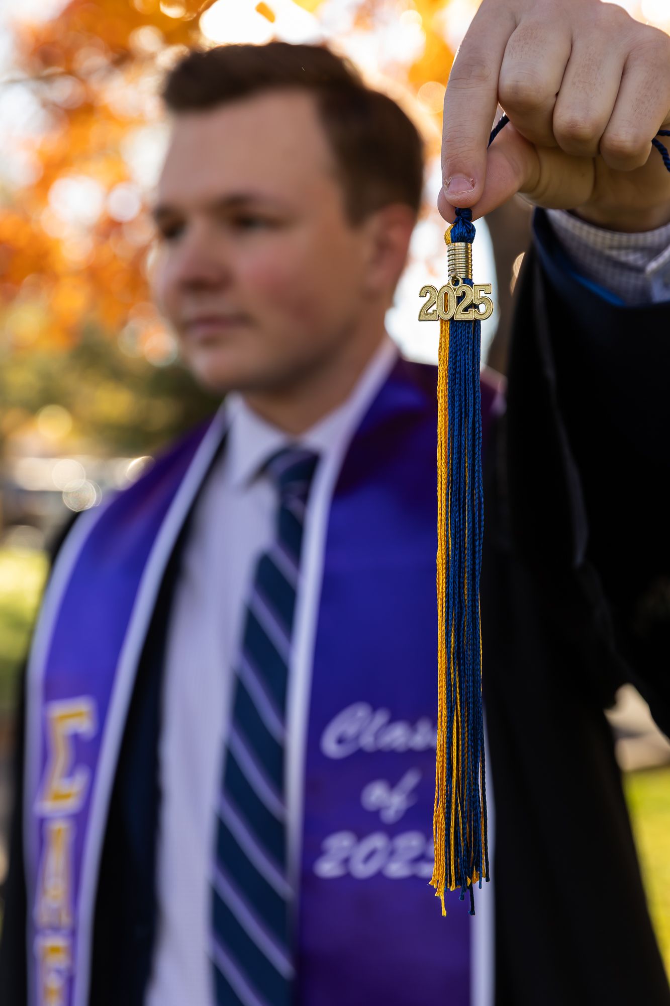 Graduates in blue and white stoles stand in line during outdoor commencement ceremony with autumn foliage in background.