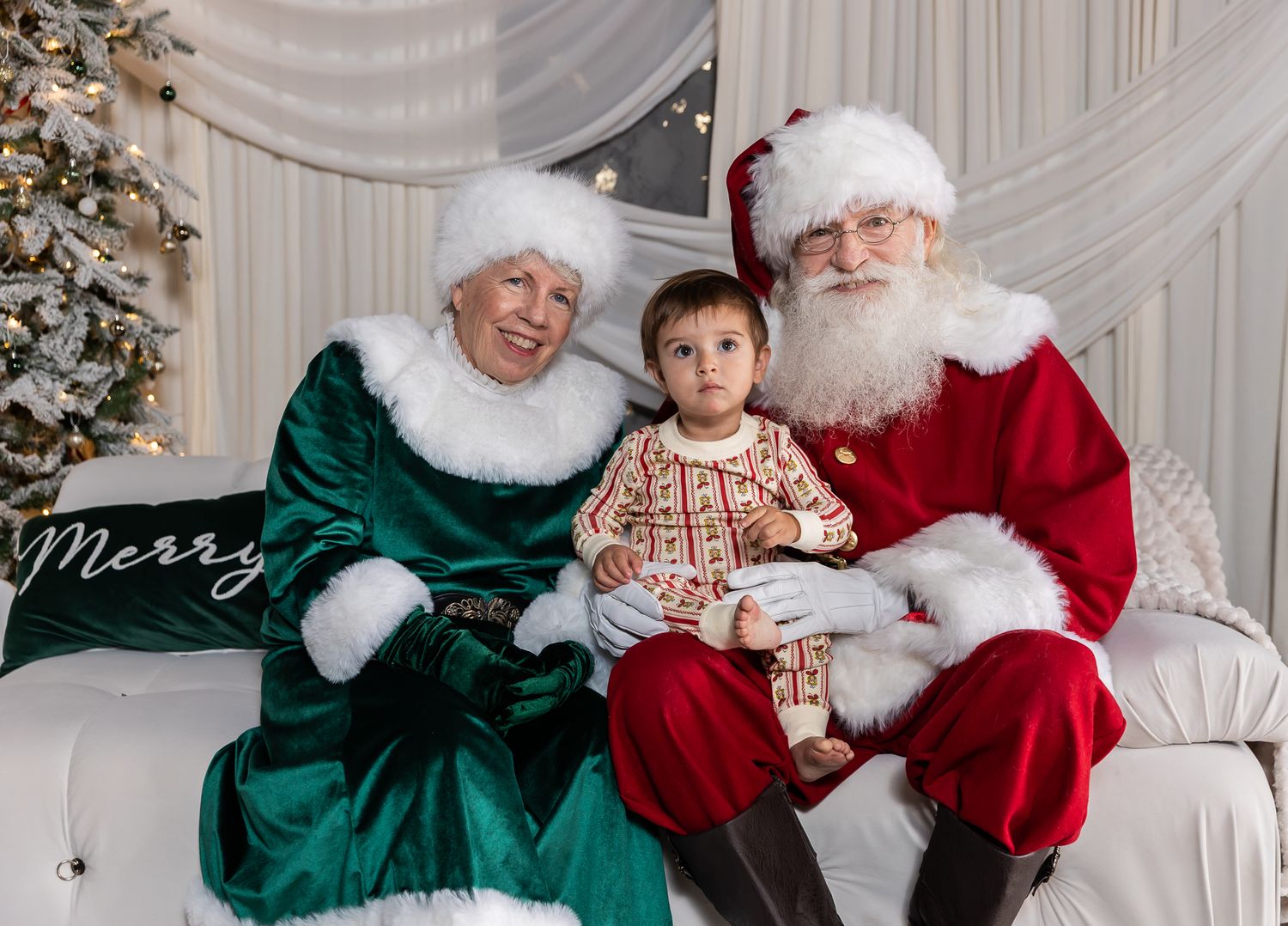 A toddler sits on Santa's lap beside Mrs. Claus in a green velvet dress during a cozy Christmas photo session near a decorated tree.