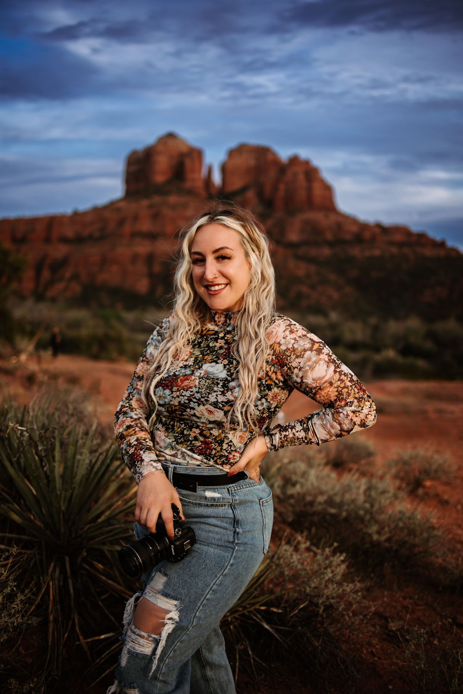 A person in a floral top and ripped jeans poses against the dramatic red rock formations of Sedona at sunset.