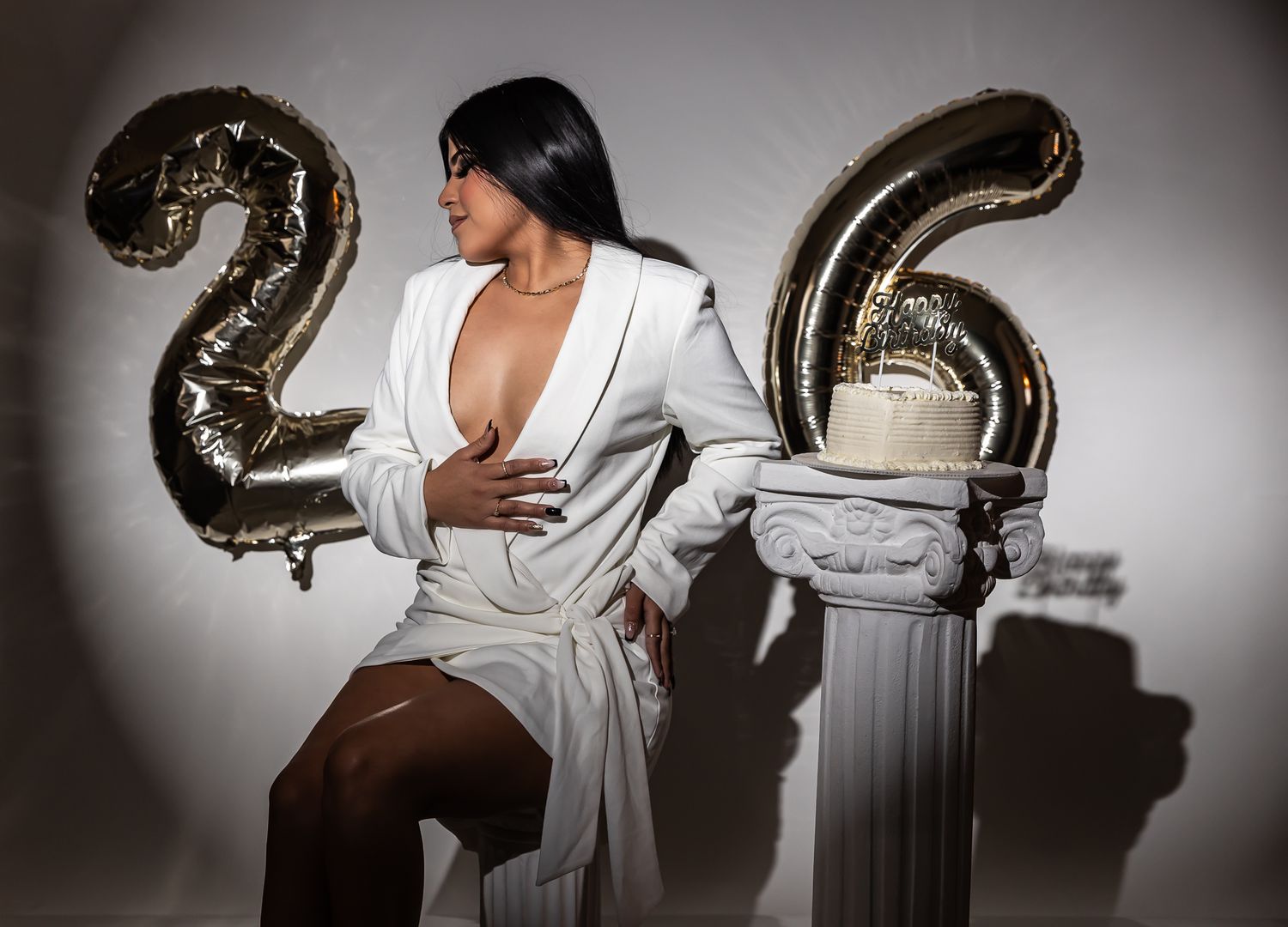 A person in a white robe poses against a dark background with metallic balloon numbers for a birthday celebration.