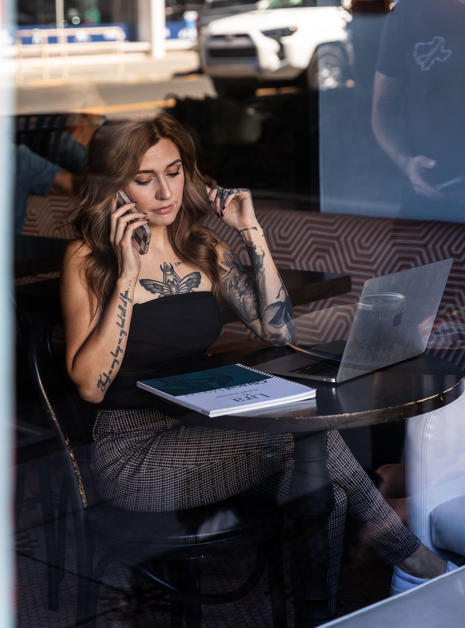 Tattooed woman multitasking on phone and laptop at cafe table, wearing black top and plaid pants.