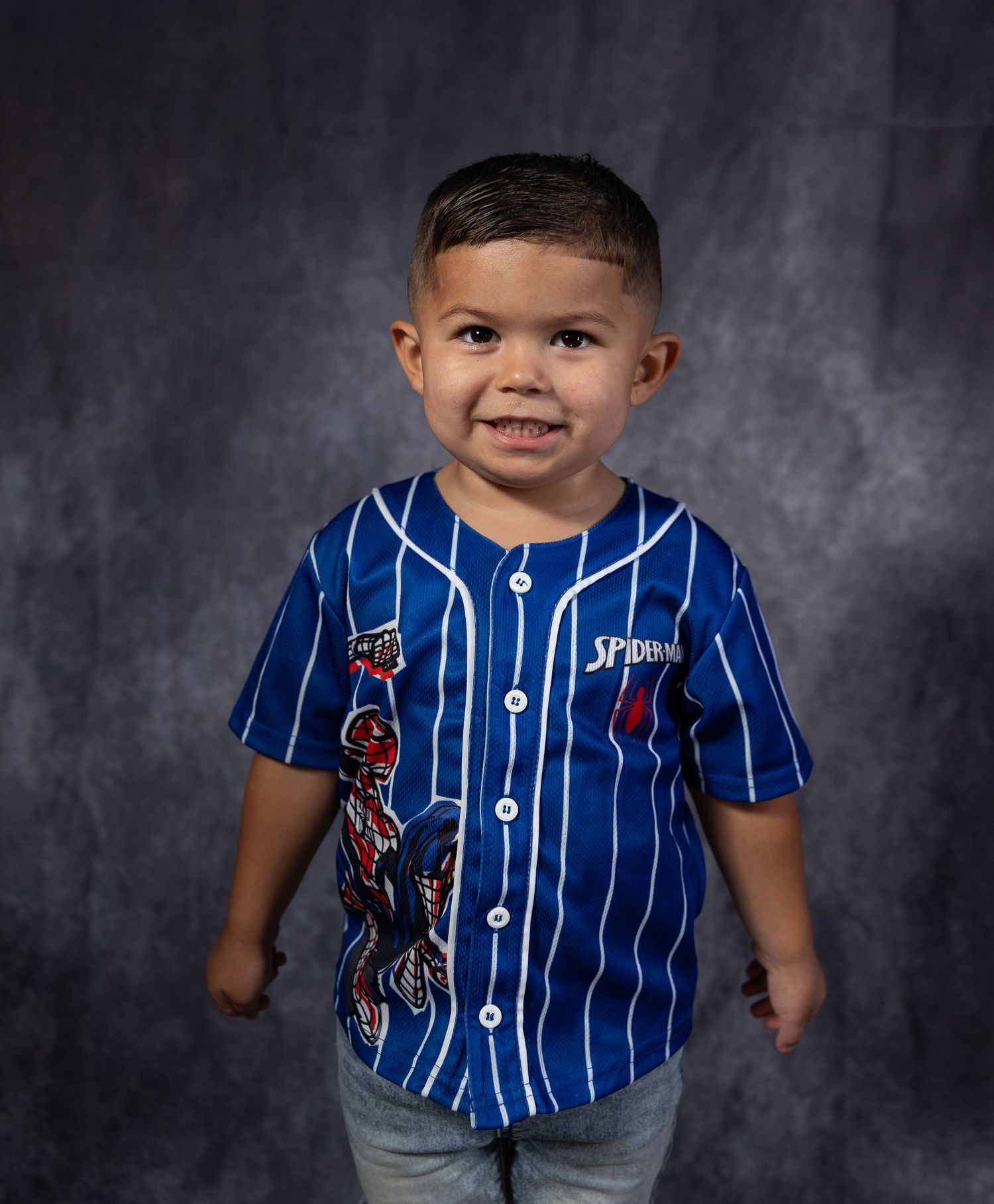 Young boy smiling in a blue pinstripe Spider-Man baseball jersey, posing for a portrait photo.
