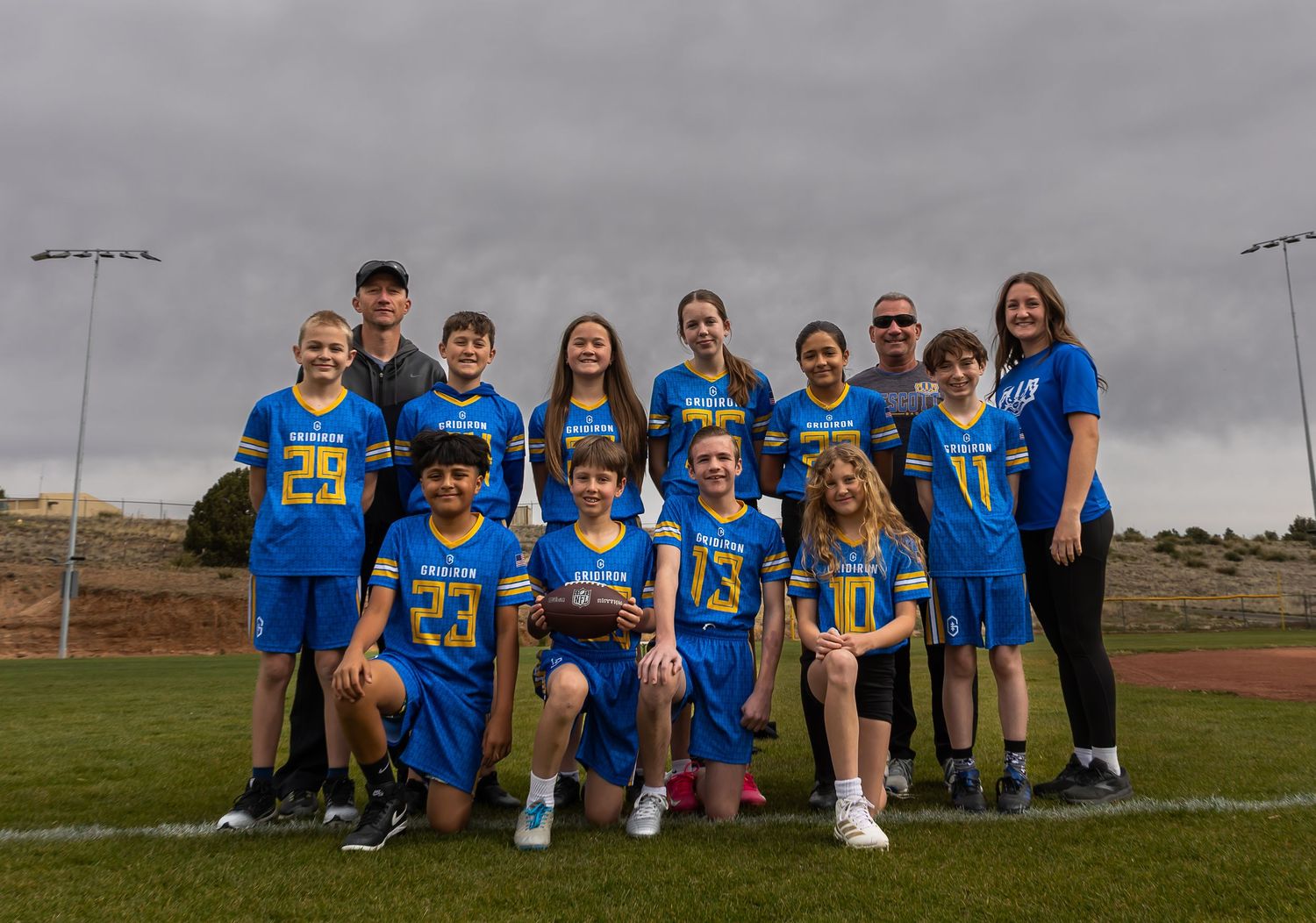 Youth lacrosse team in blue and gold uniforms posing together on a sports field under cloudy skies.