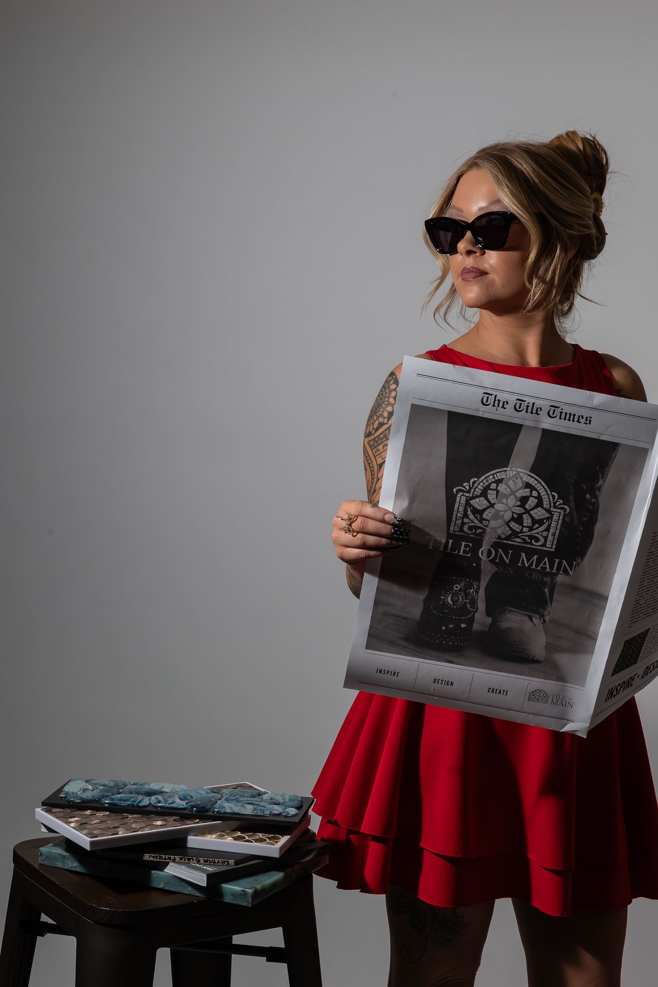 Woman in red dress and sunglasses holding The Tile Times newspaper, posing beside a stool with magazines in a studio.