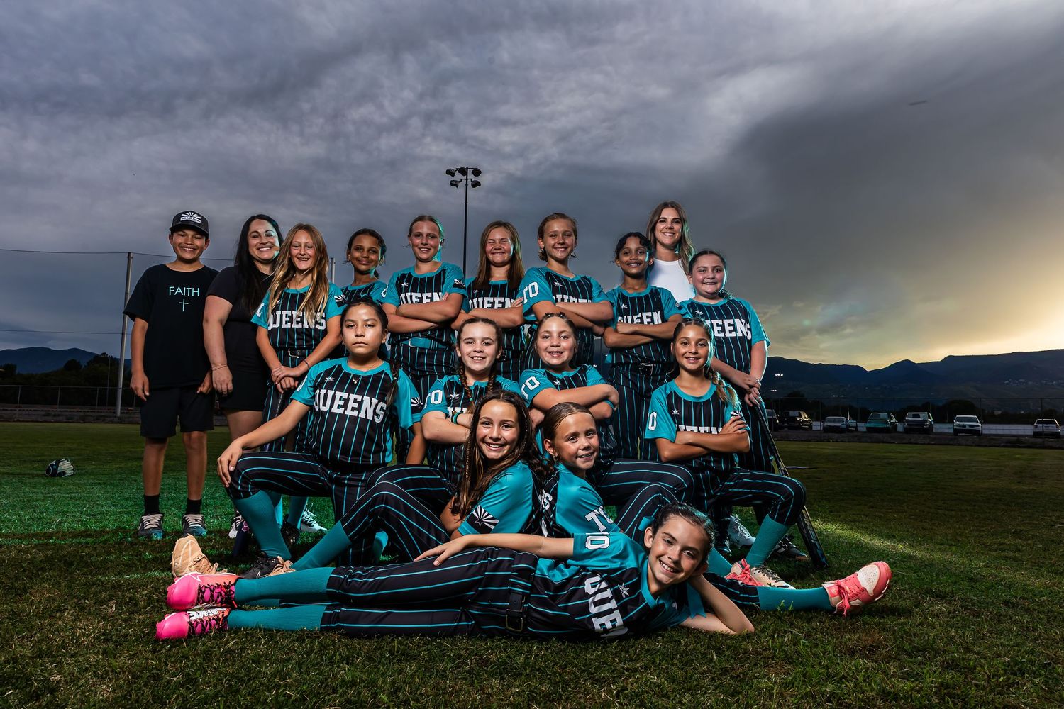 Girls softball team posing together on field wearing teal Queens uniforms under dramatic stormy sky.