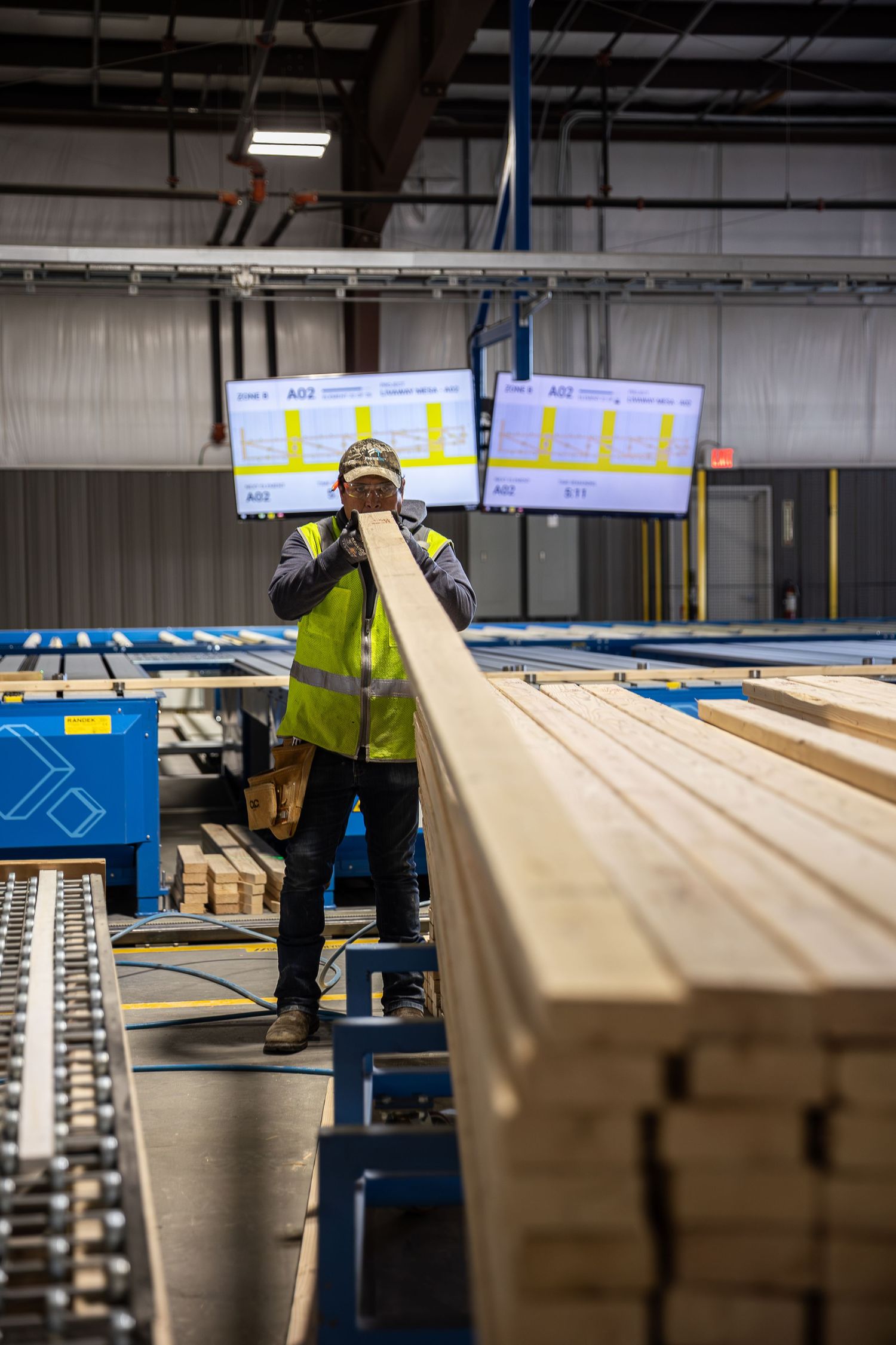 Worker in safety vest inspects lumber at an industrial wood processing facility with digital displays in background.
