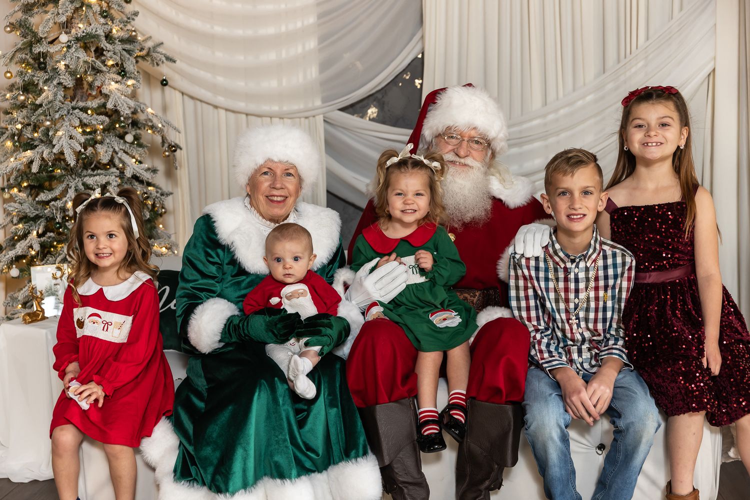 Santa Claus and Mrs. Claus pose with five smiling children in festive holiday outfits beside a decorated Christmas tree.