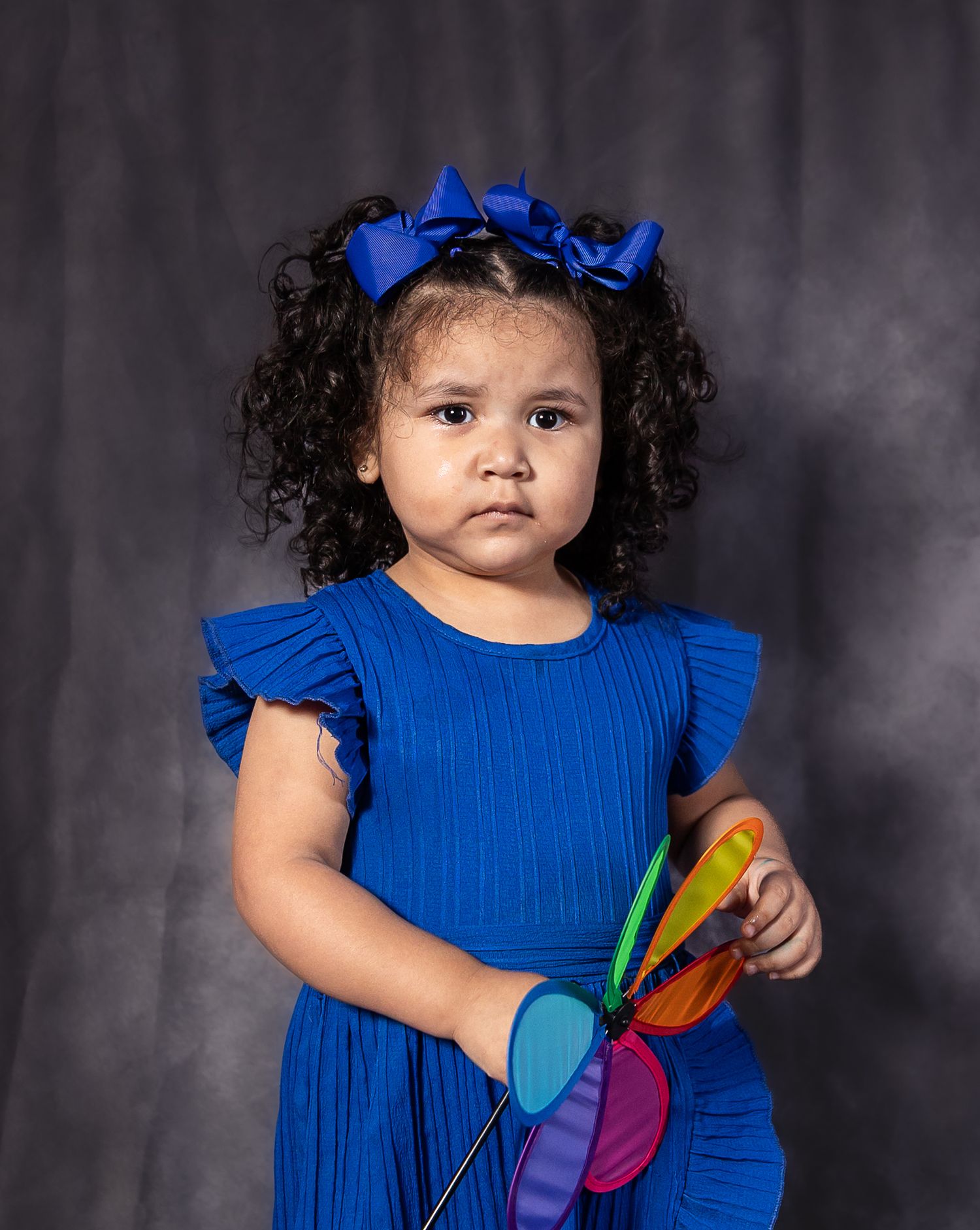 Young girl in blue dress and bow holds colorful pinwheel toy, posing for portrait photo.