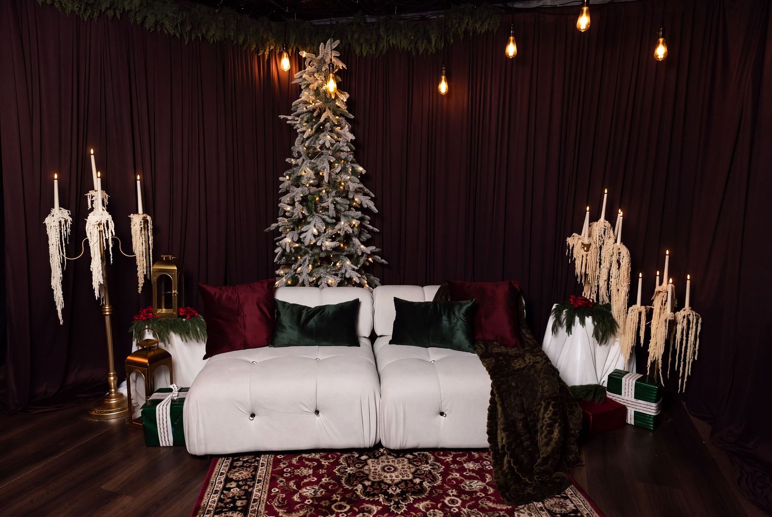 Cozy holiday photo studio set with a flocked Christmas tree, white tufted sofa, dark green and burgundy pillows, and a vintage red rug.