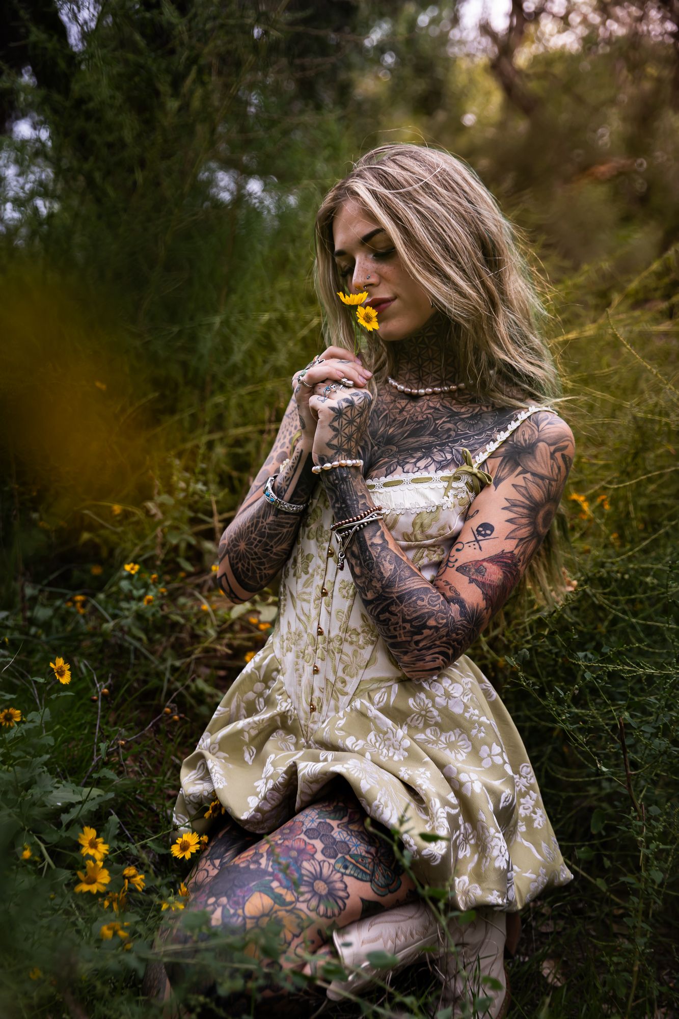 A person wearing a floral lace dress poses gracefully in a lush garden setting with yellow wildflowers and greenery.