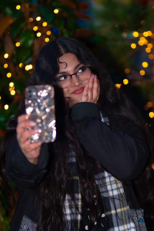 Young woman with long dark hair and glasses taking a selfie at night with bokeh lights behind her.
