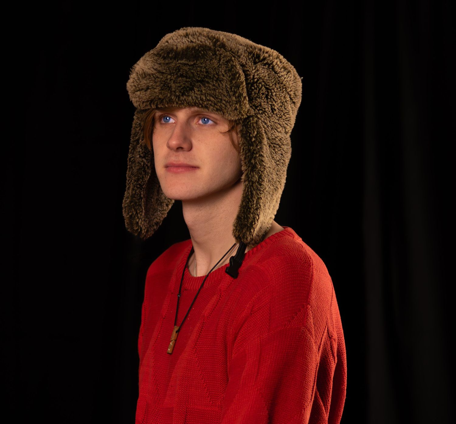 Young man wearing a brown fur trapper hat and red sweater posing against a dark background.