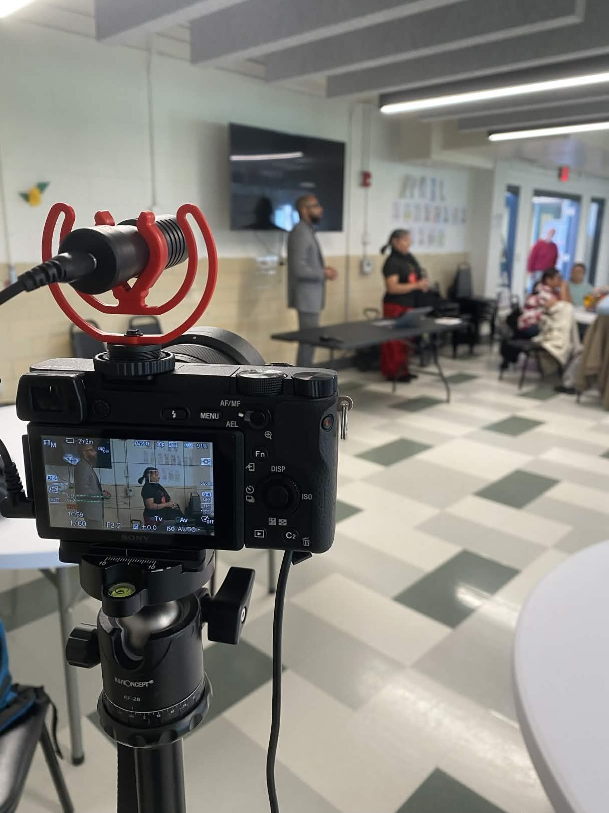 Camera on tripod recording a presenter speaking to a small group in a community meeting room.
