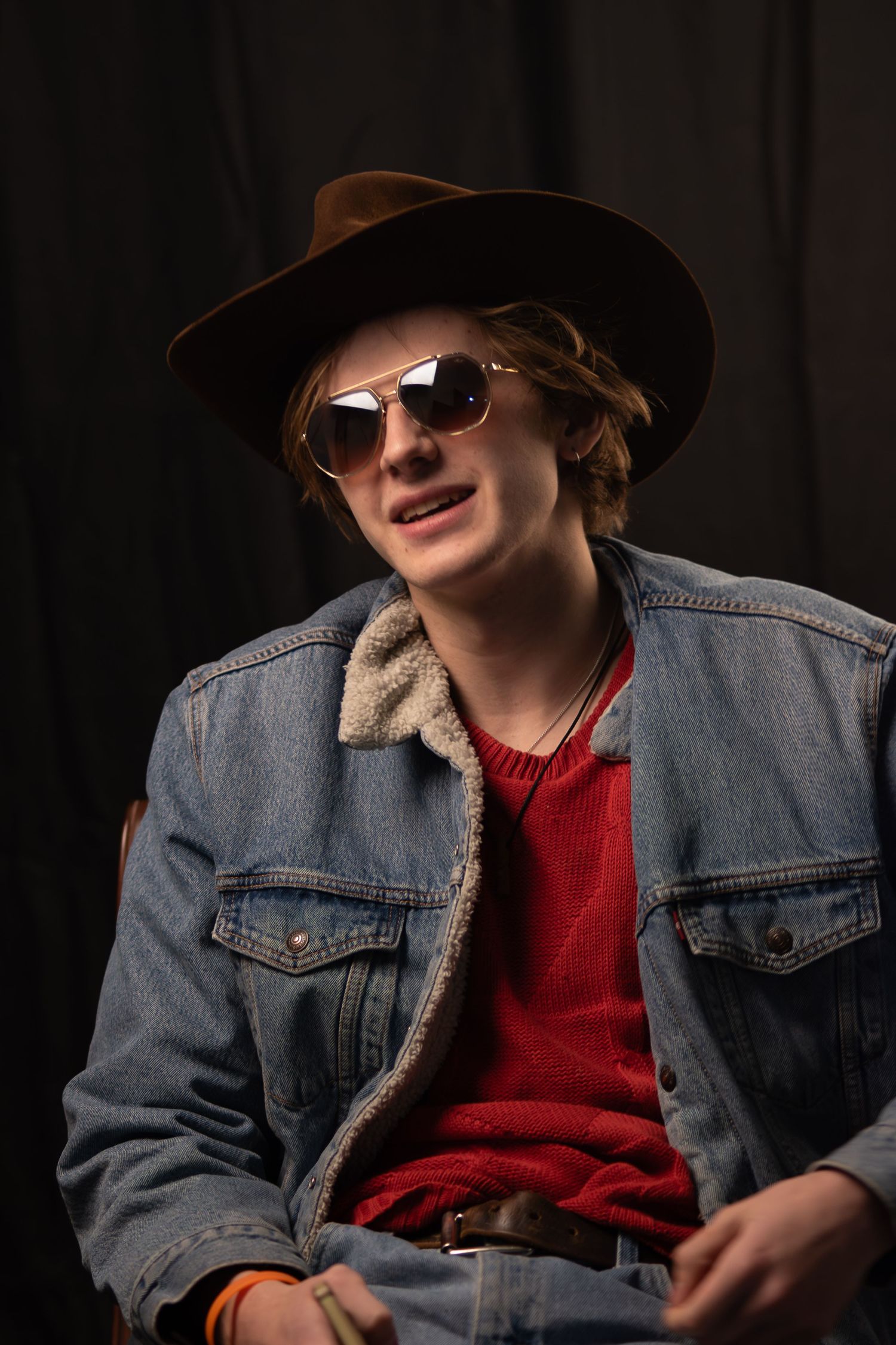Young man wearing a brown fedora, sunglasses, and denim jacket sitting against a dark background.