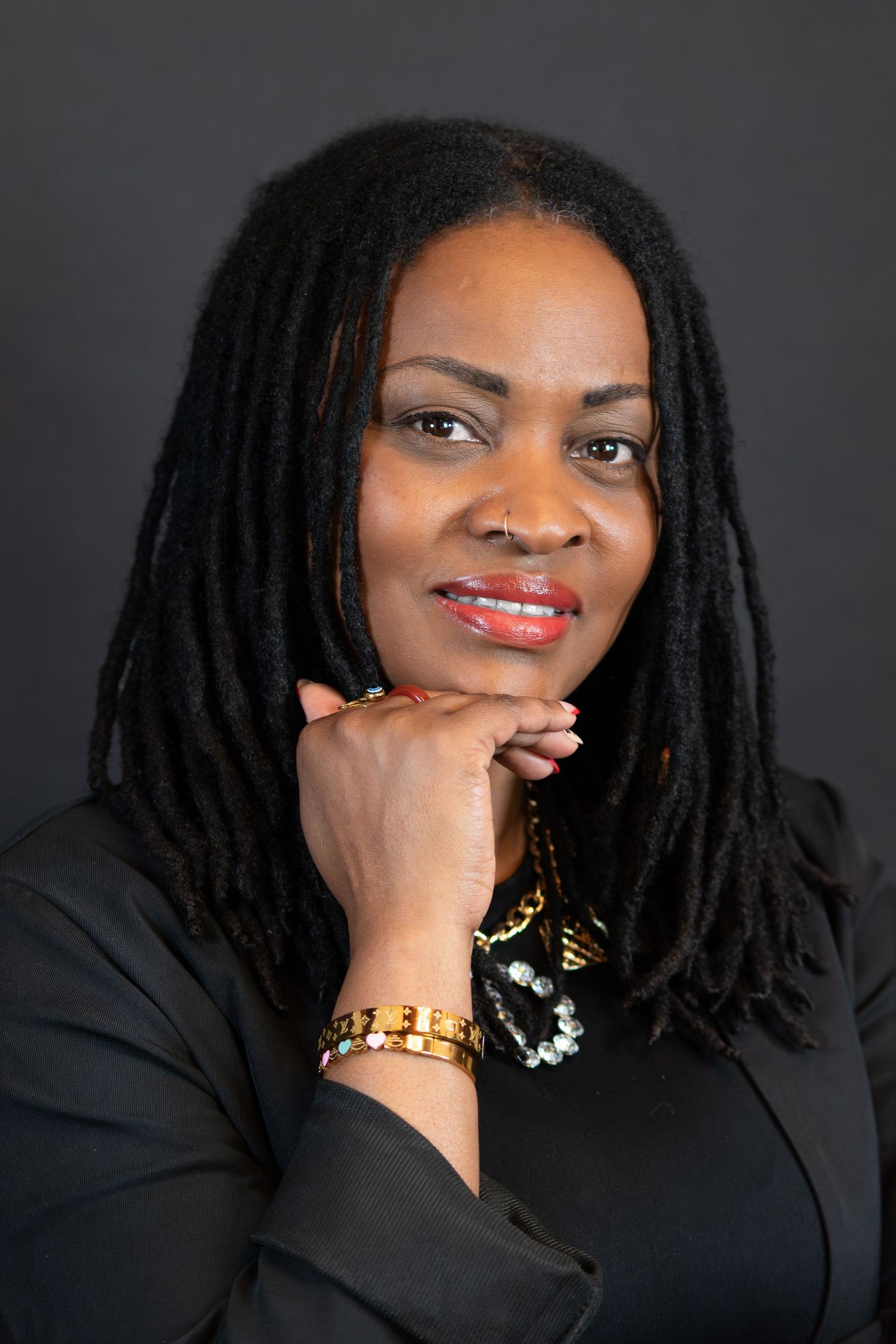 Professional portrait of a woman with long dreadlocks wearing a black blazer and gold bracelet, hands posed under chin.