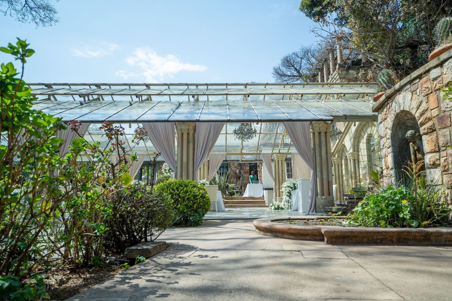 Elegant glass greenhouse wedding venue with draped curtains, stone walls, and lush garden surroundings on a sunny day.
