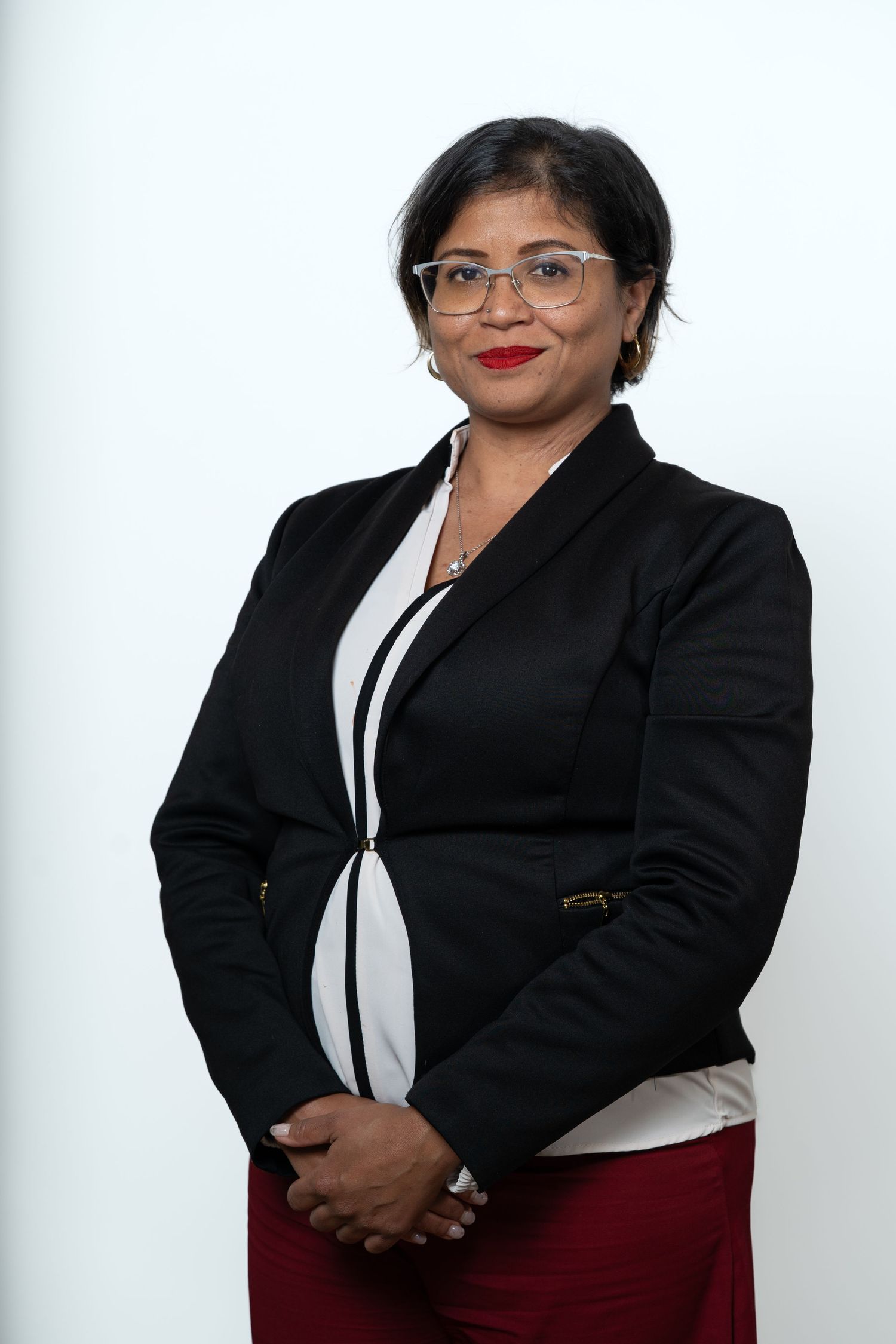 Professional woman in black blazer and red pants, wearing glasses, posing confidently against white background.