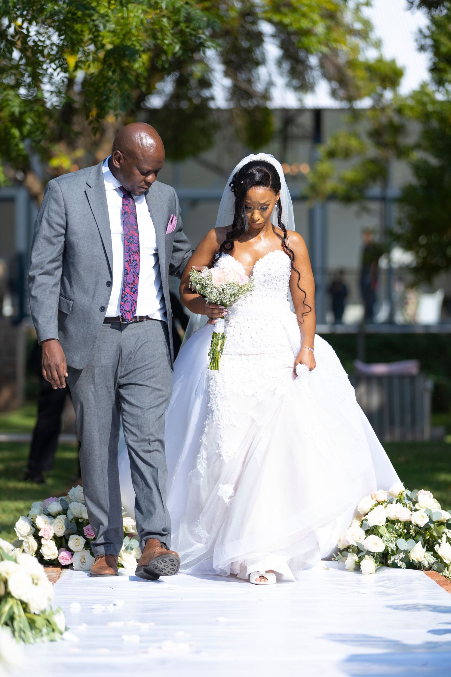 Bride in a white lace ballgown walks the aisle with escort during an outdoor wedding ceremony.