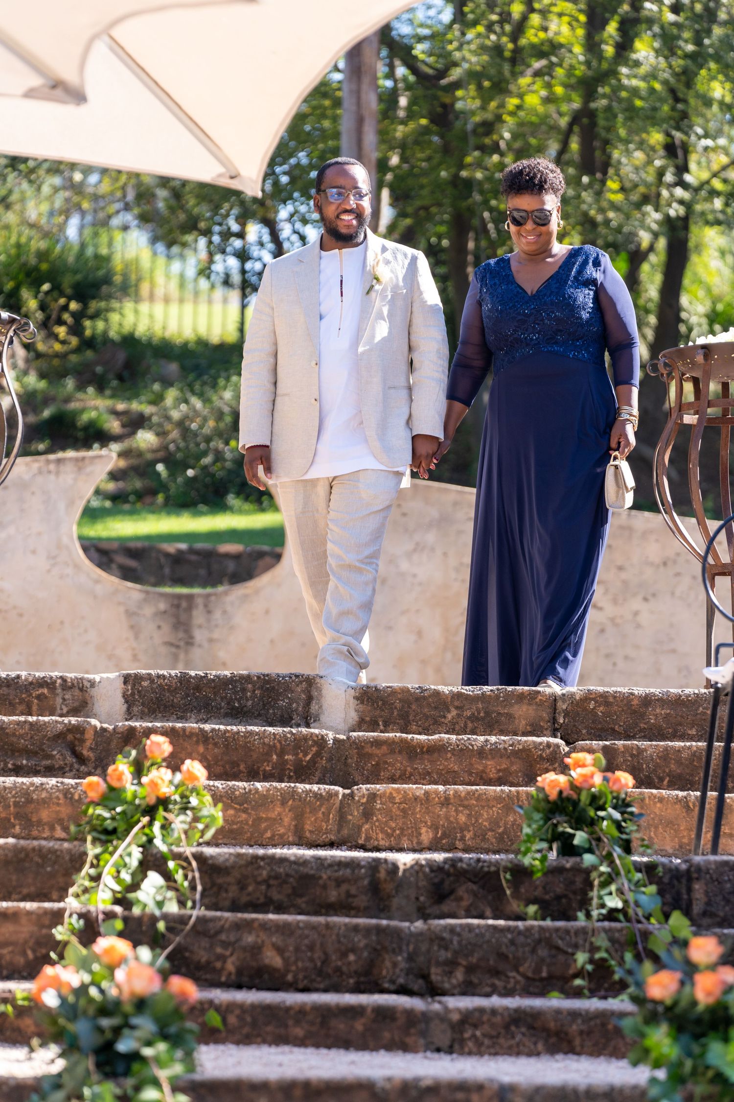 Groom in beige suit and woman in navy blue dress descending stone steps at outdoor wedding ceremony.