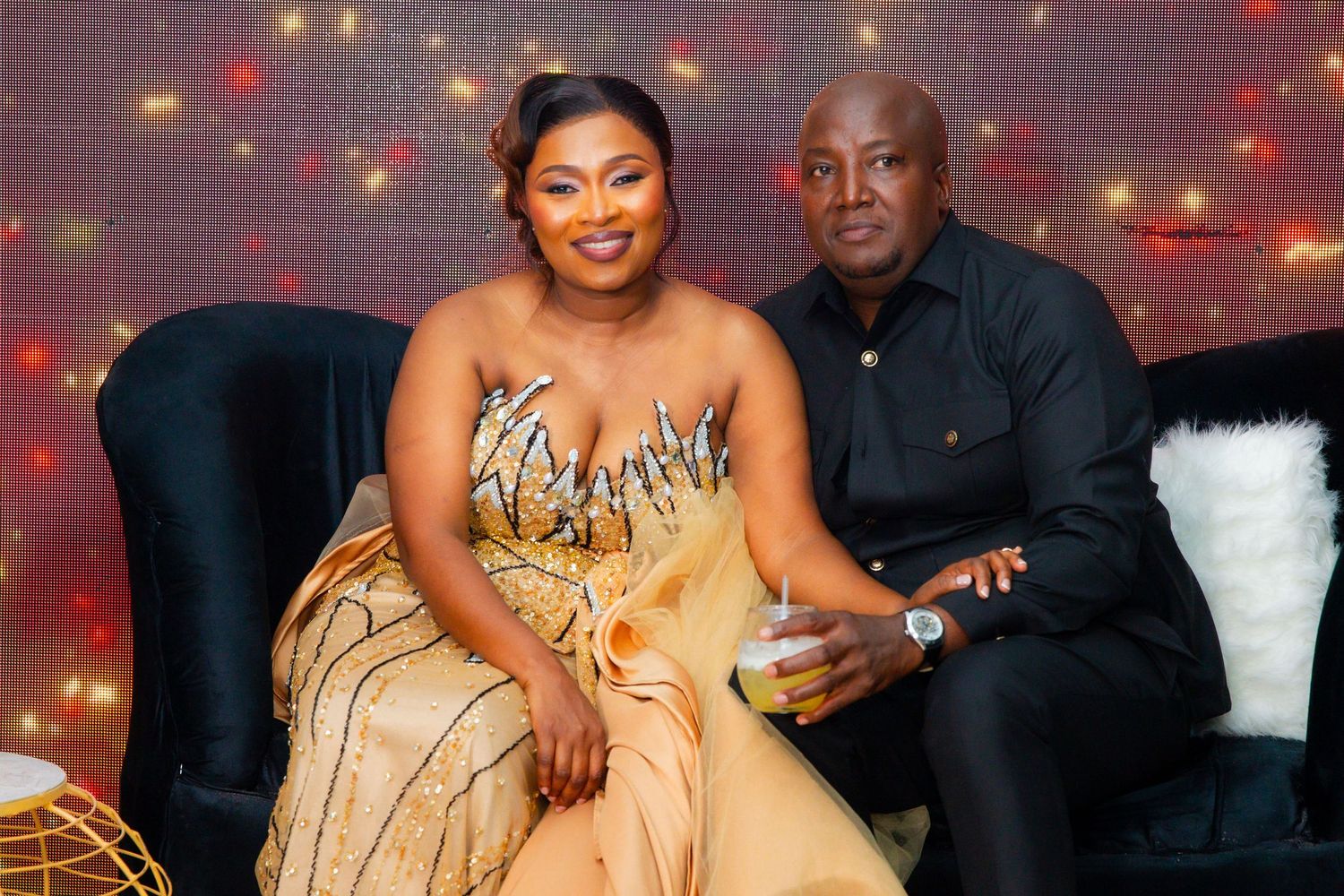 Couple posing on a velvet sofa in gold and black formal attire against a glittering backdrop.