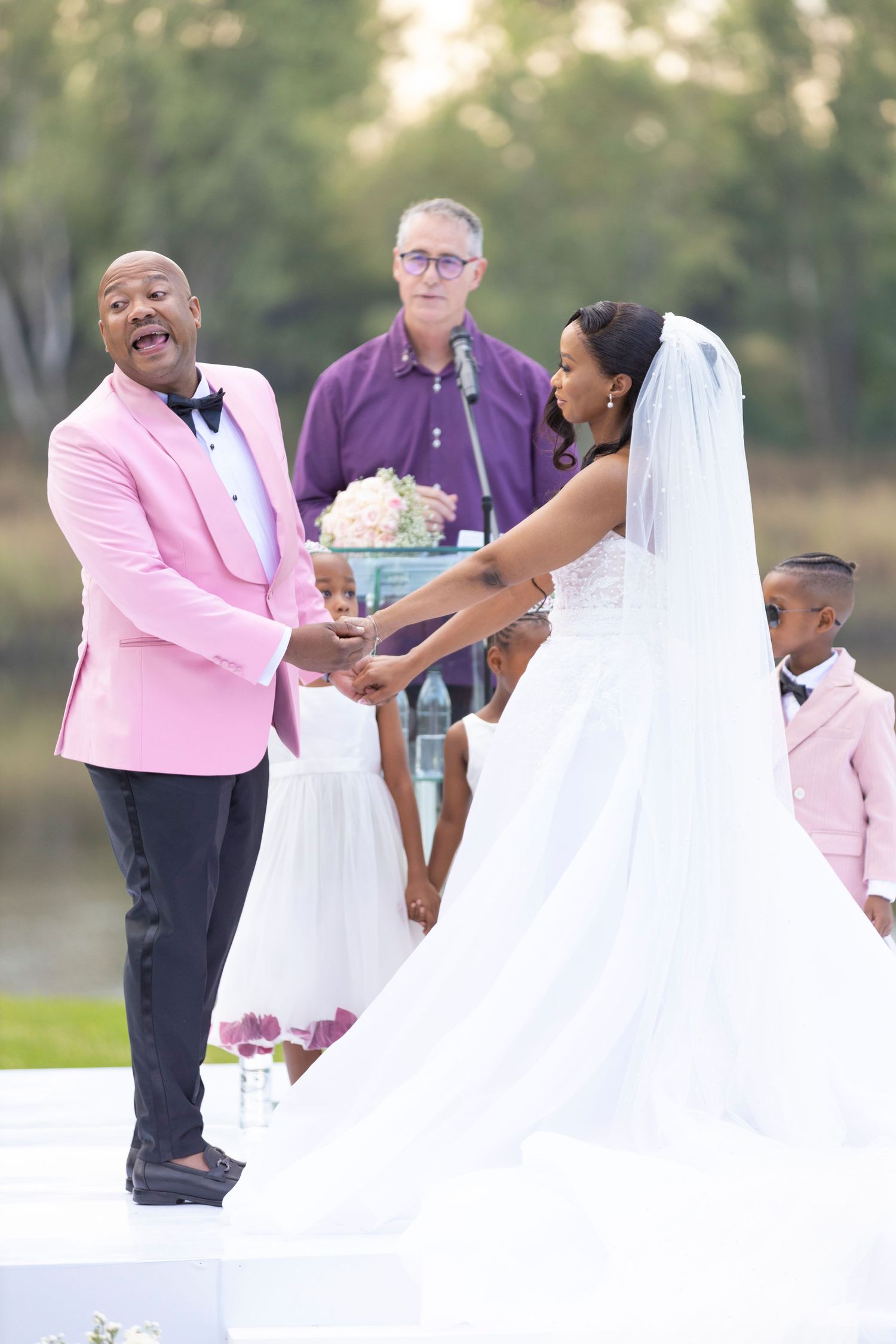 Groom in pink tuxedo reacts emotionally while holding bride's hands during outdoor wedding ceremony.