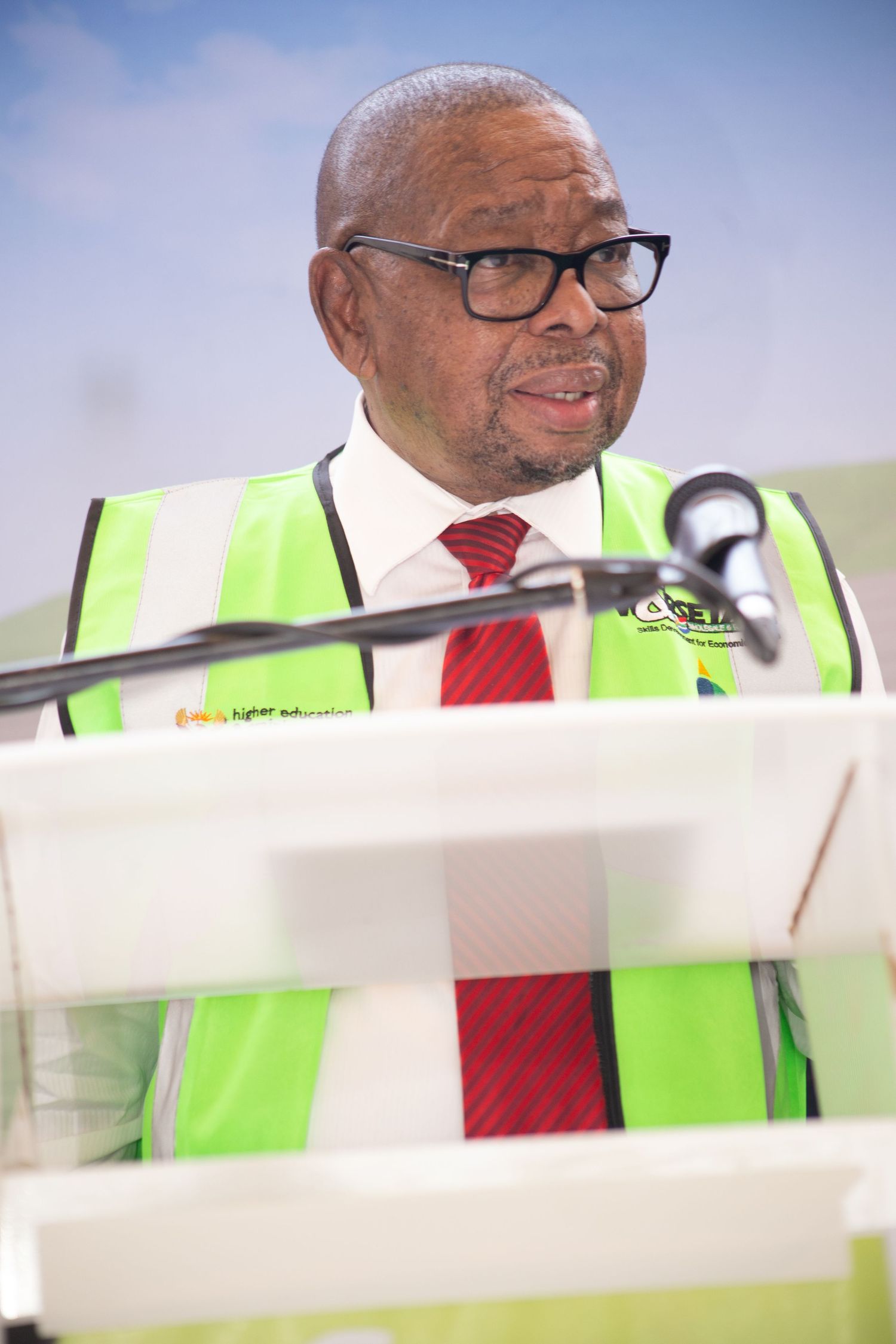 An older man wearing a green safety vest and red tie speaks at a podium during an outdoor event.
