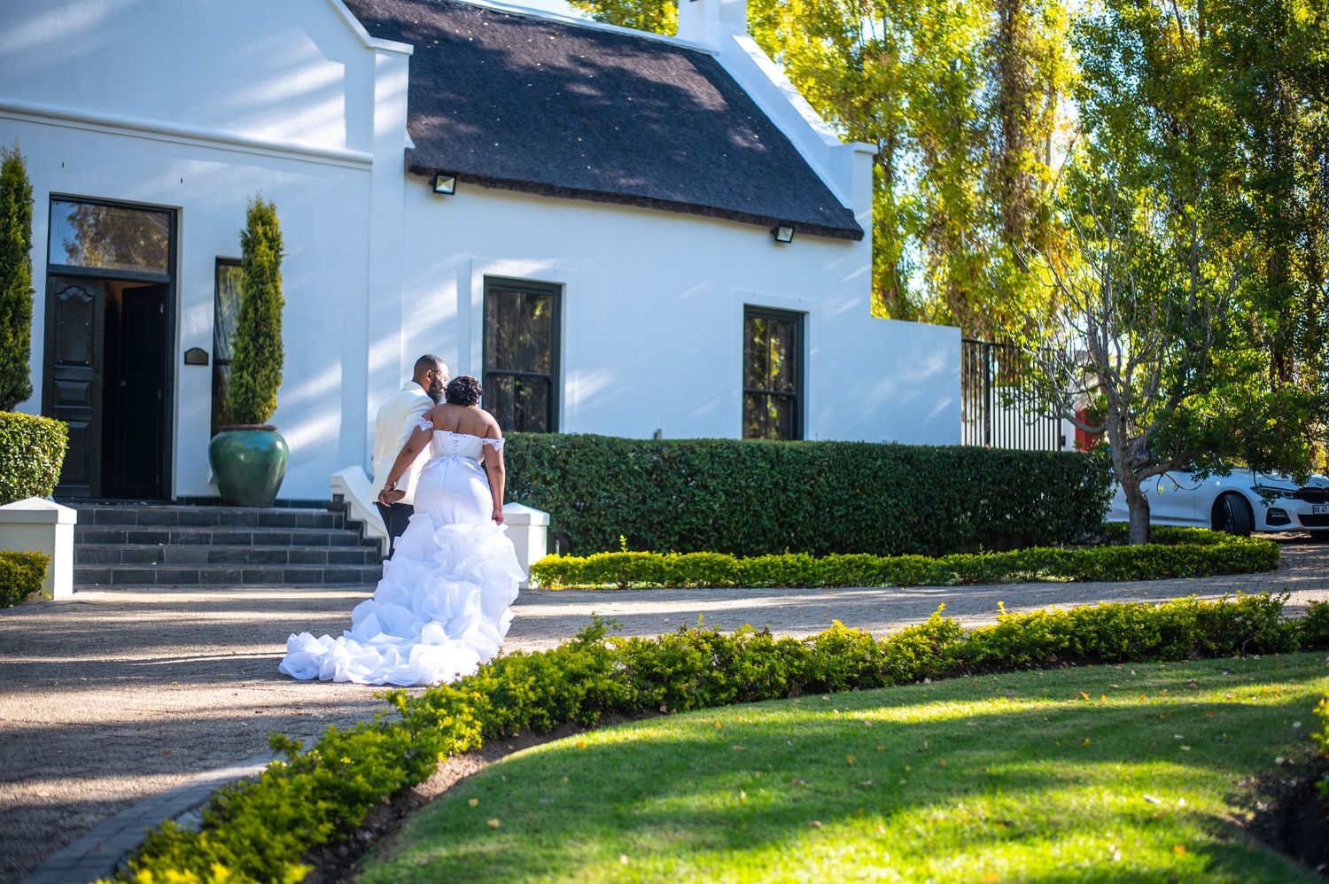Bride in stunning ruffled mermaid wedding dress walking along garden path beside white Cape Dutch style venue.