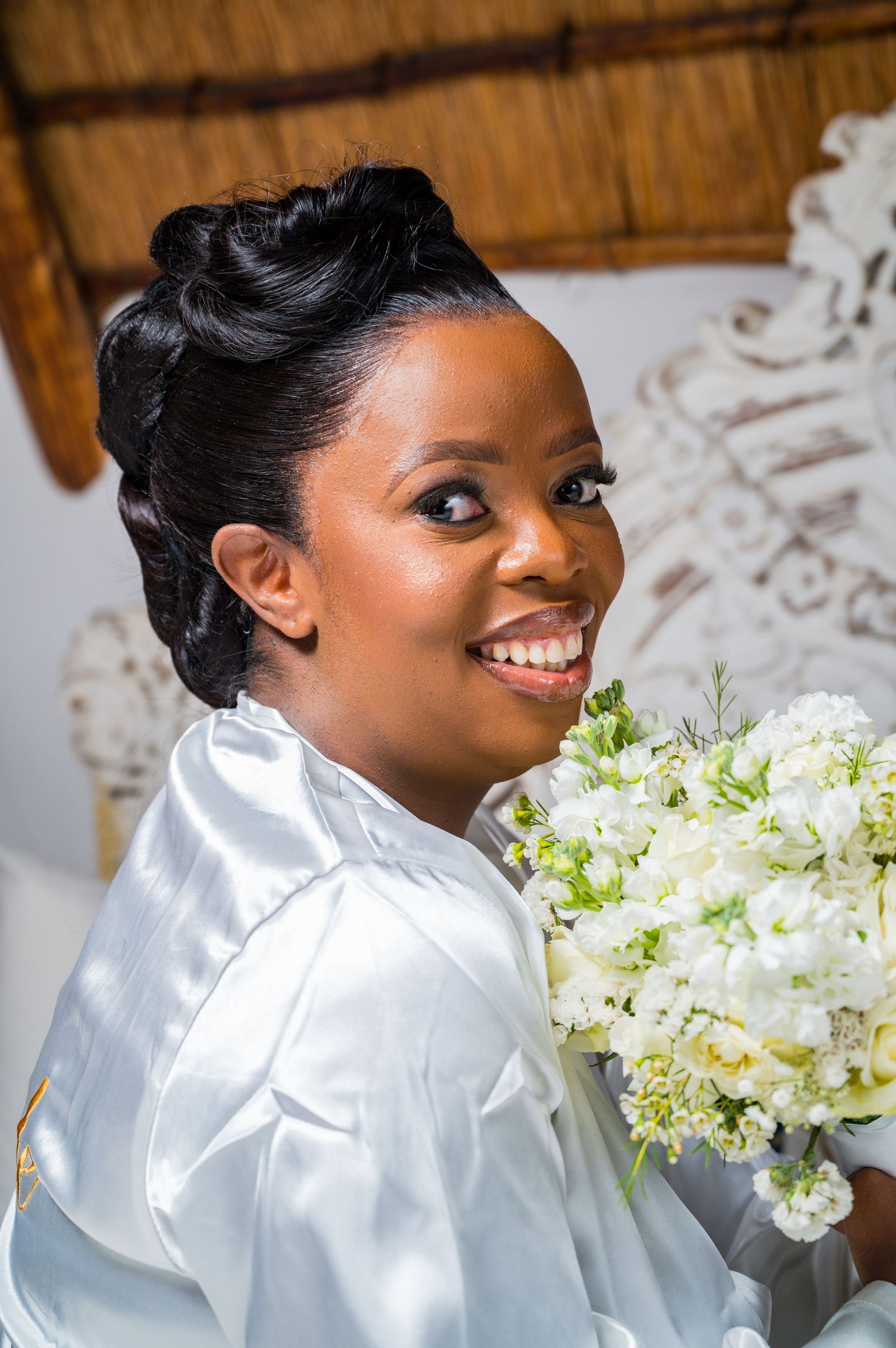 Radiant bride in white satin robe holds white floral bouquet and smiles over her shoulder.
