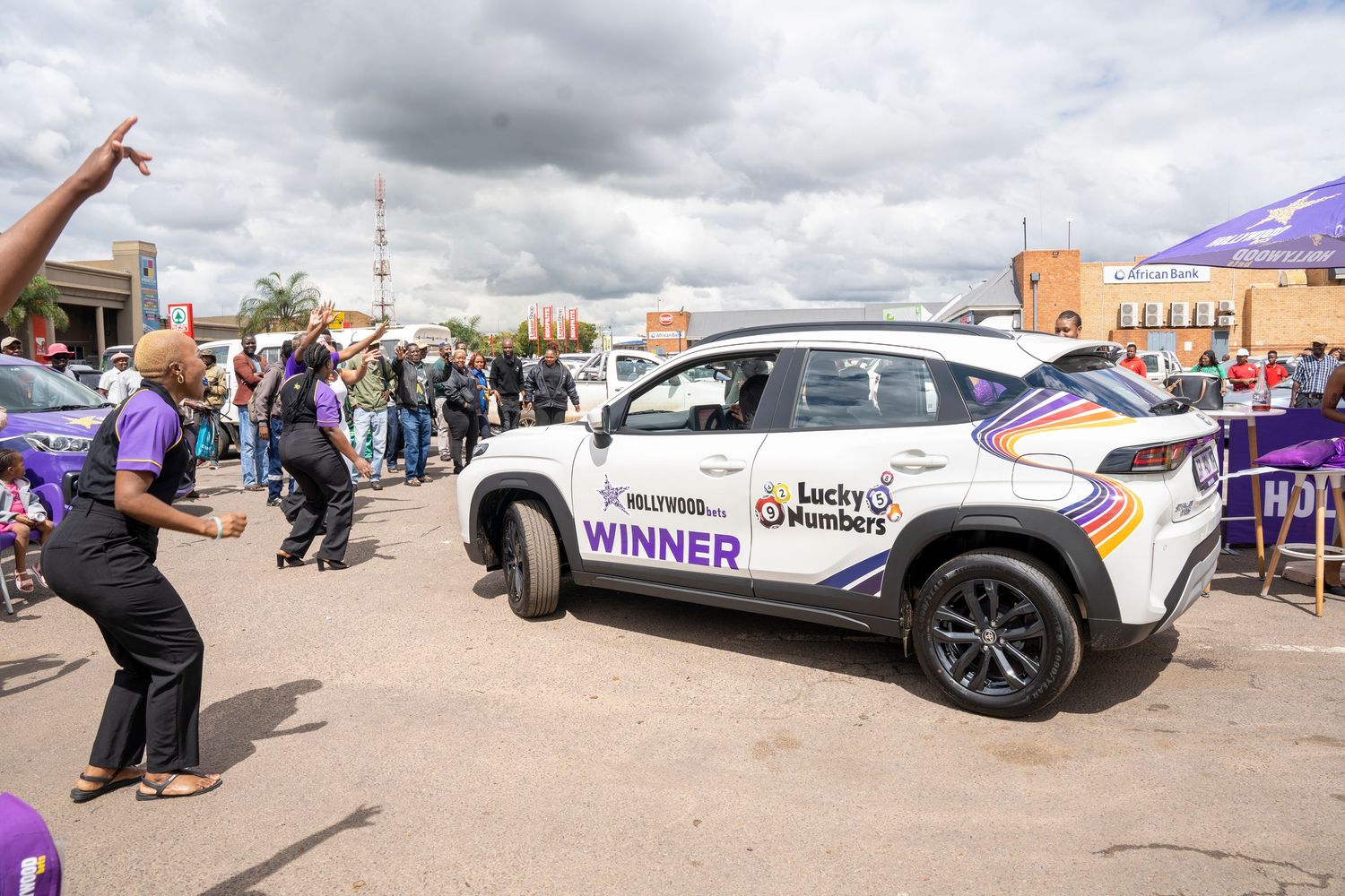 Hollywoodbets Lucky Numbers winner car on display at outdoor prize-giving event with crowd celebrating.