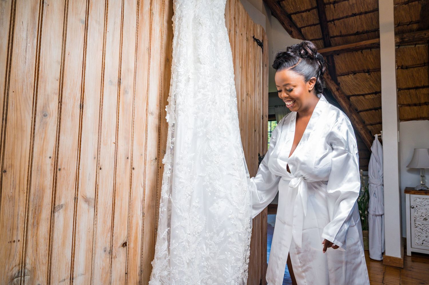 Smiling bride in white robe admiring her lace wedding dress hanging in rustic room.
