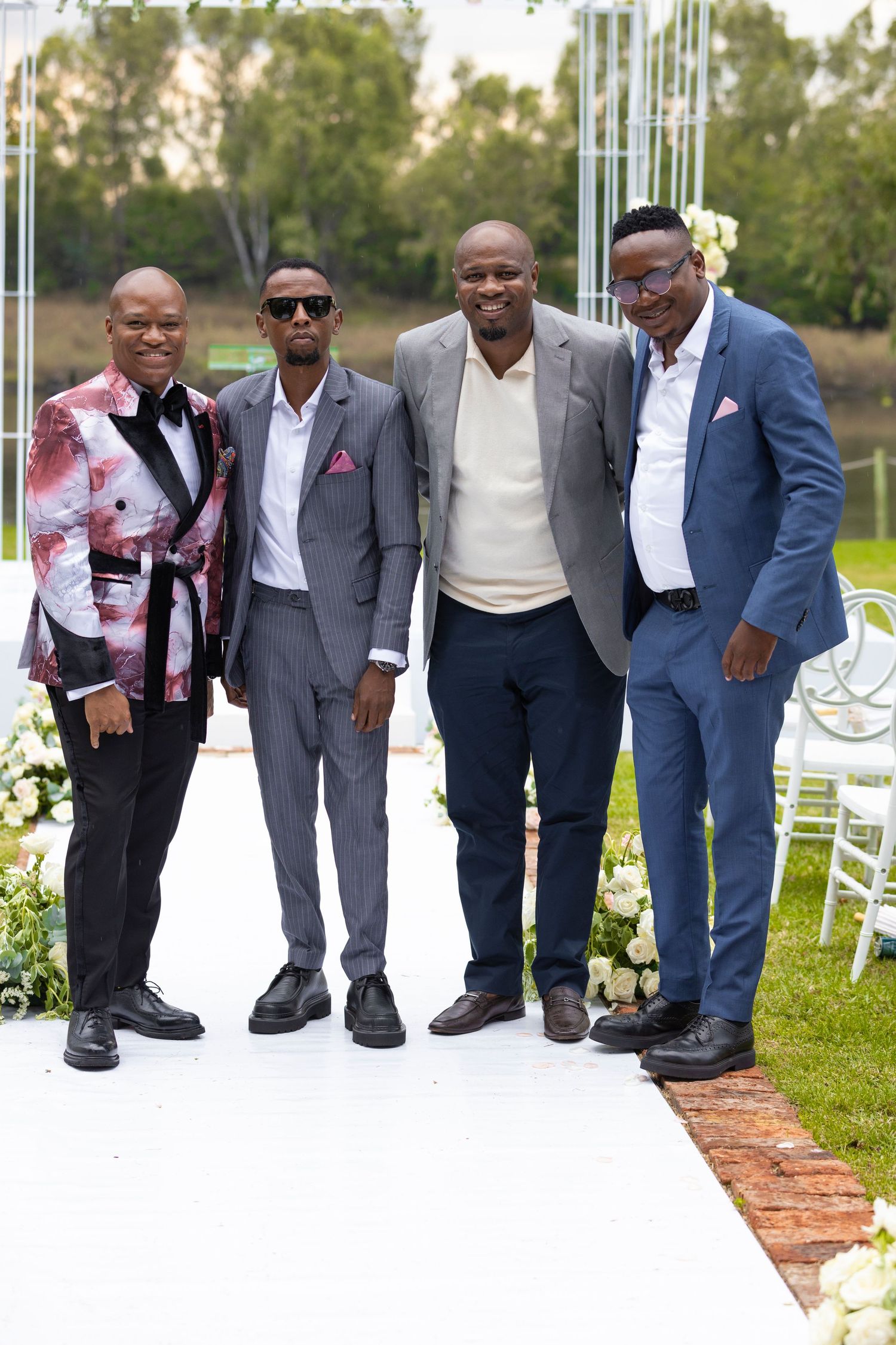 Four well-dressed men pose together on the white aisle runner at an outdoor wedding celebration.