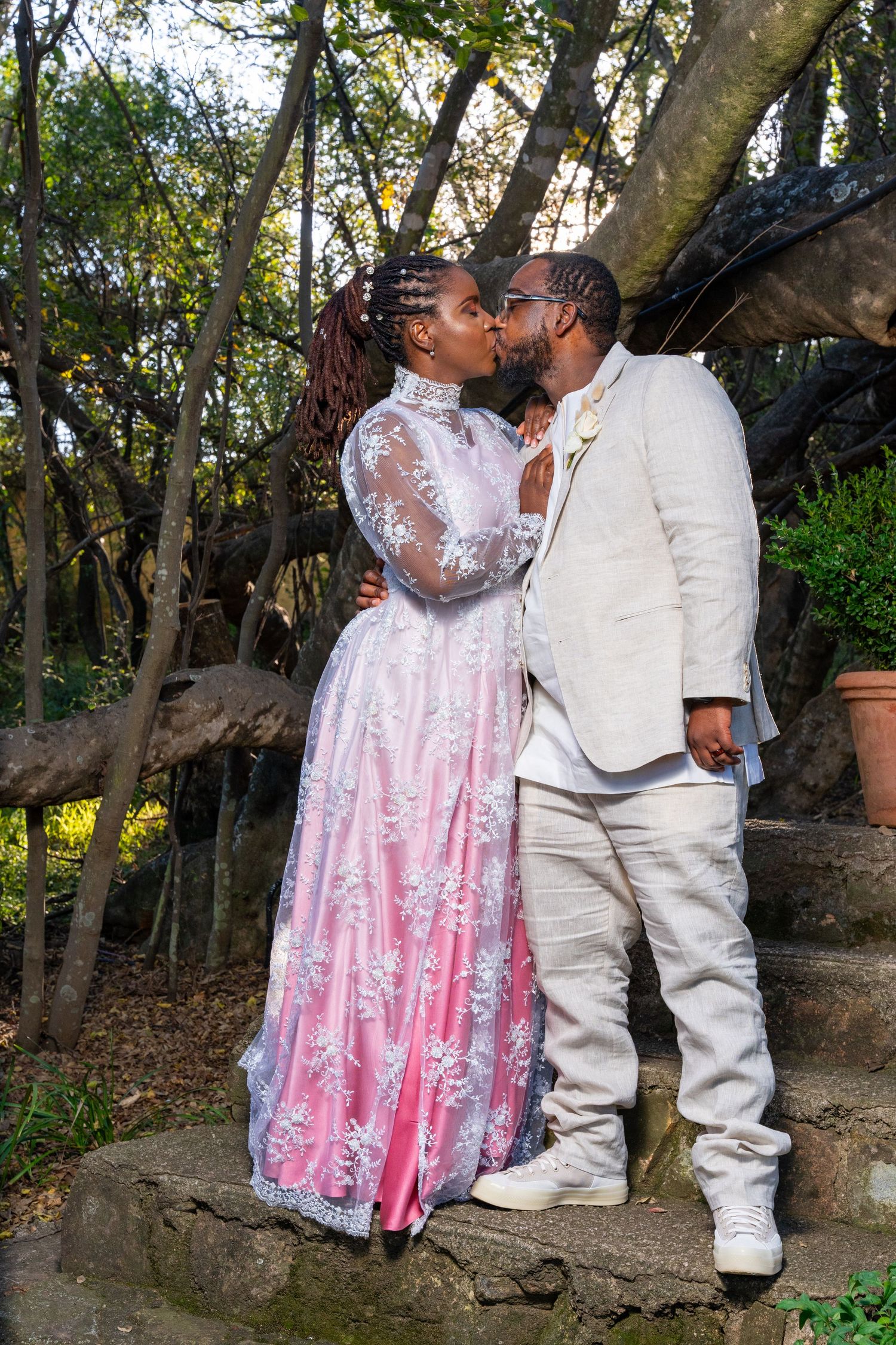 Bride and groom sharing a kiss outdoors, bride in pink and white lace gown, groom in white suit.
