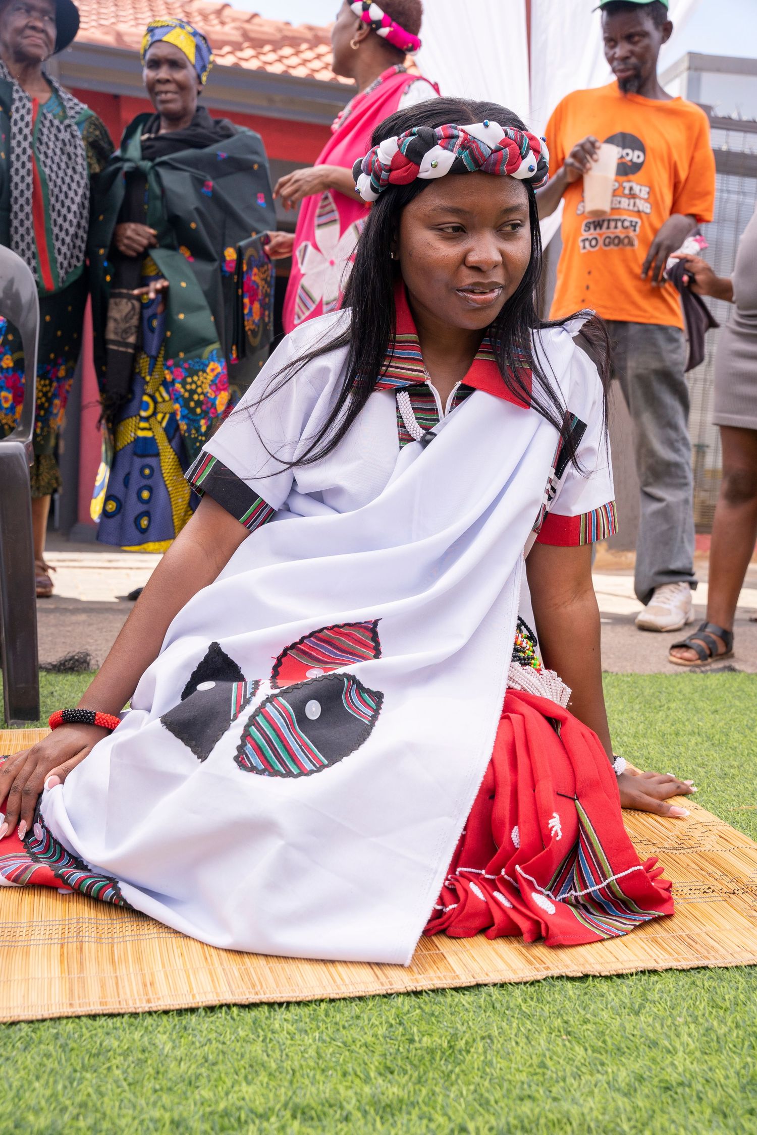 Young woman in traditional Xhosa white ceremonial dress with beaded headpiece sits gracefully on woven mat during cultural ceremony.