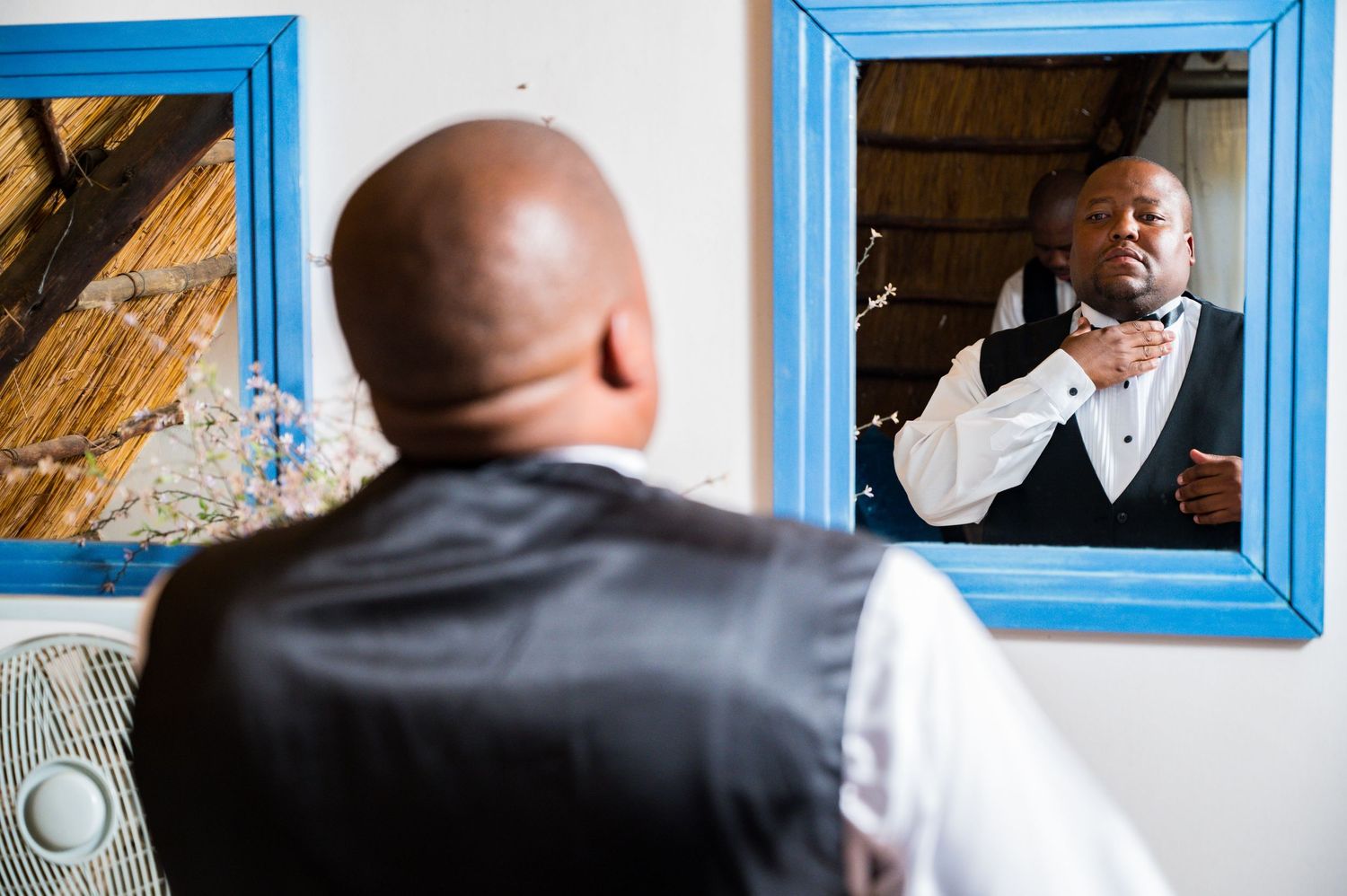 Groom adjusting bow tie while looking at reflection in blue-framed mirror before wedding.