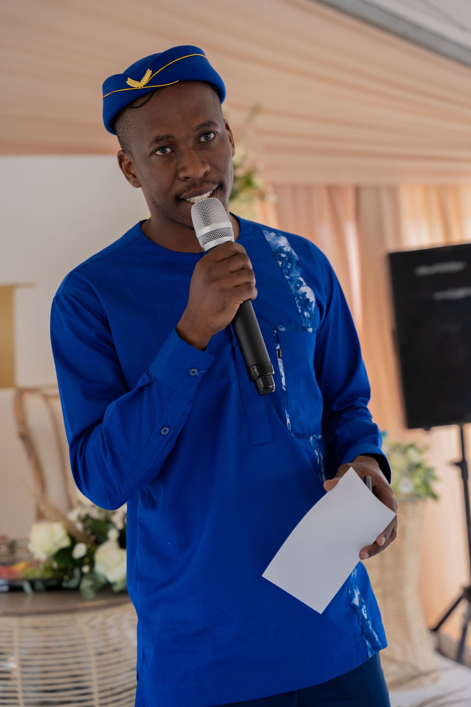 Young man in blue African traditional attire speaking into microphone at wedding reception event.