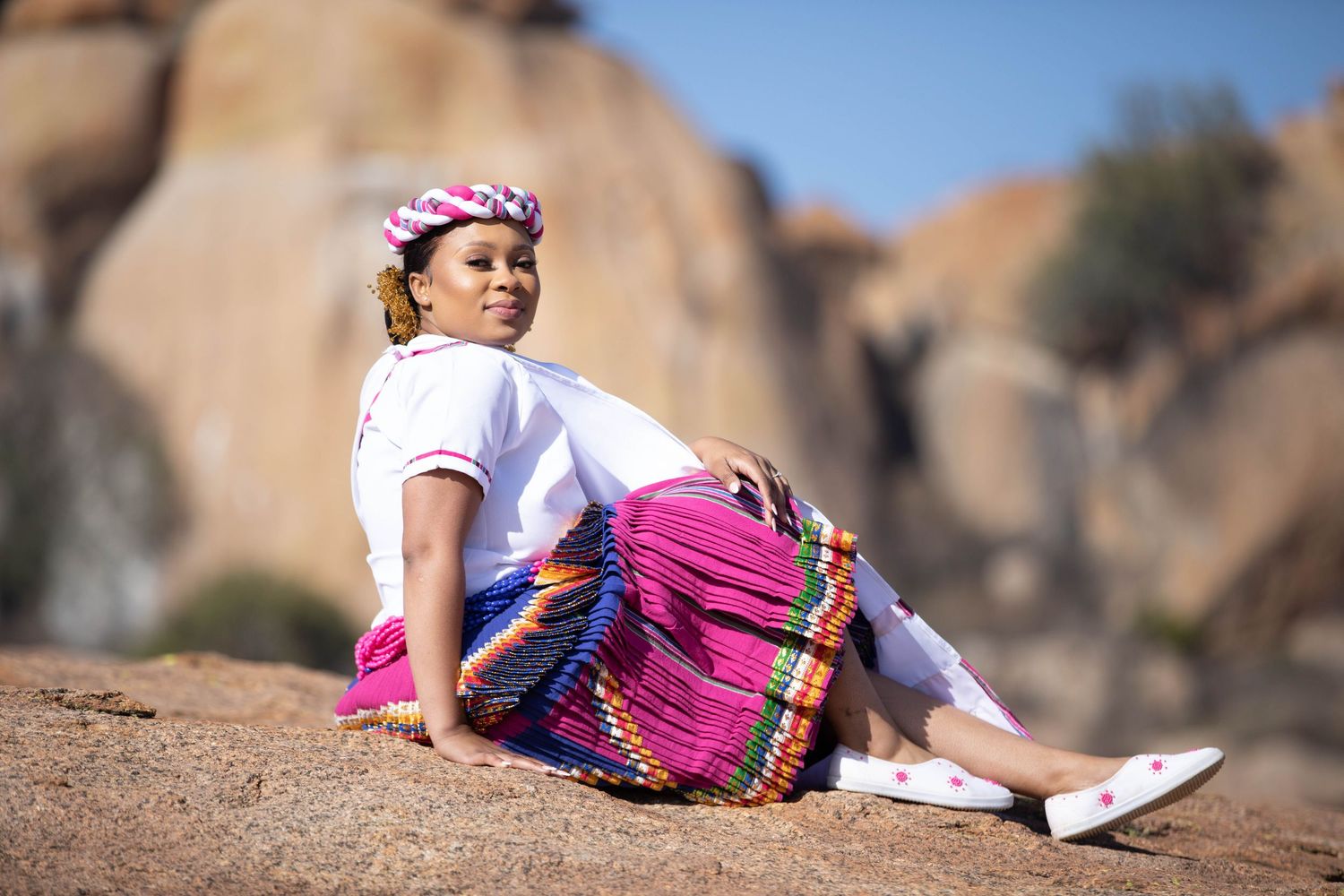 Woman in colorful traditional African attire with pink skirt and head wrap sitting on rocks outdoors.