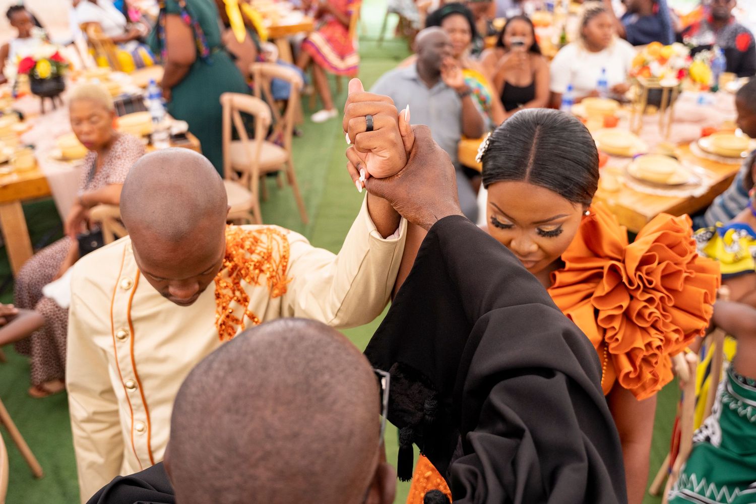 People in colorful orange attire holding hands at a festive African celebration event outdoors.