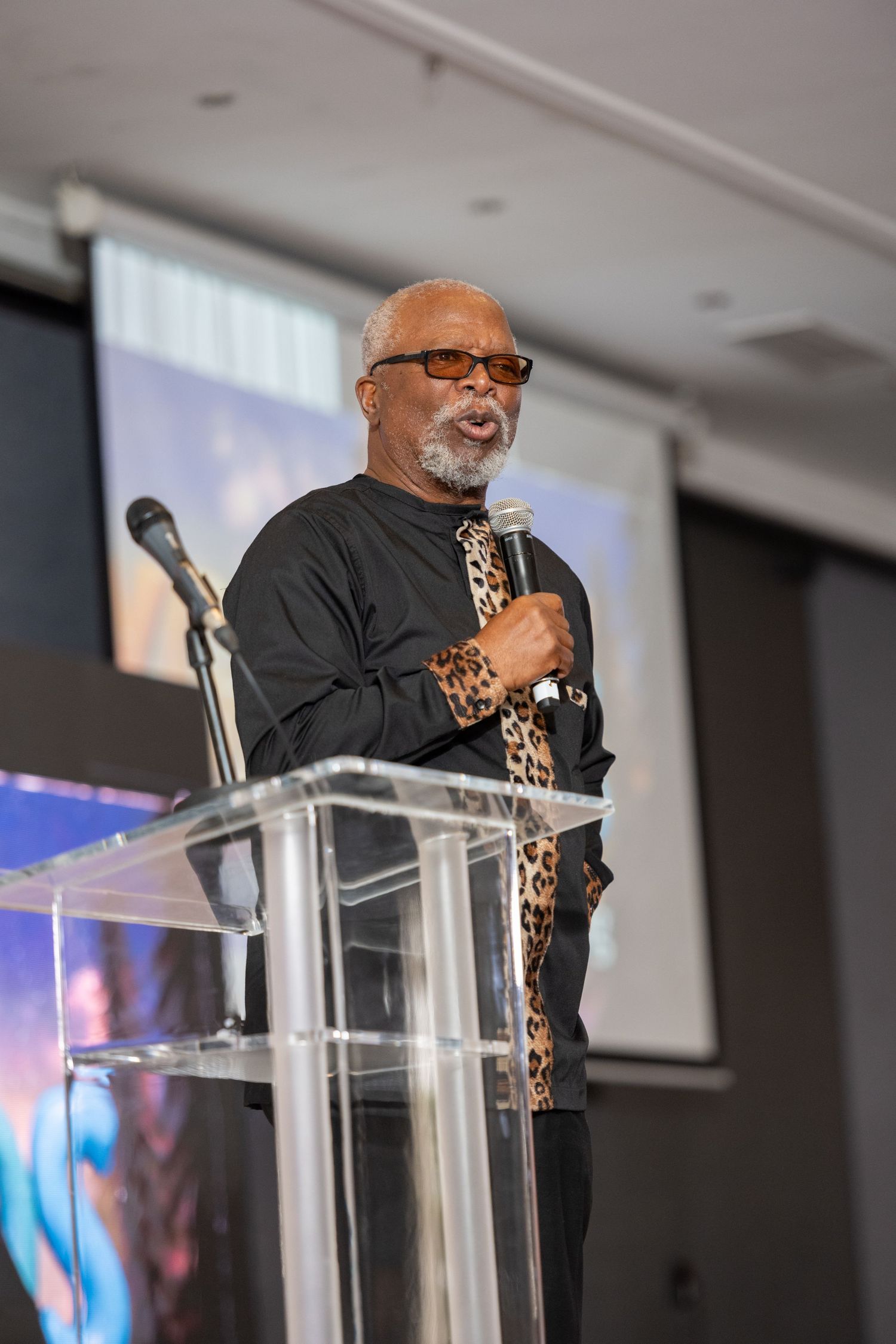 Elderly man in leopard print shirt speaking at clear podium with microphone.