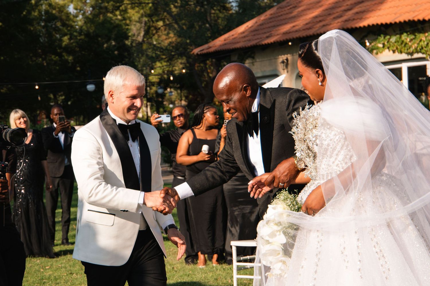Groom in white tuxedo shakes hands with guest as bride arrives at outdoor wedding.