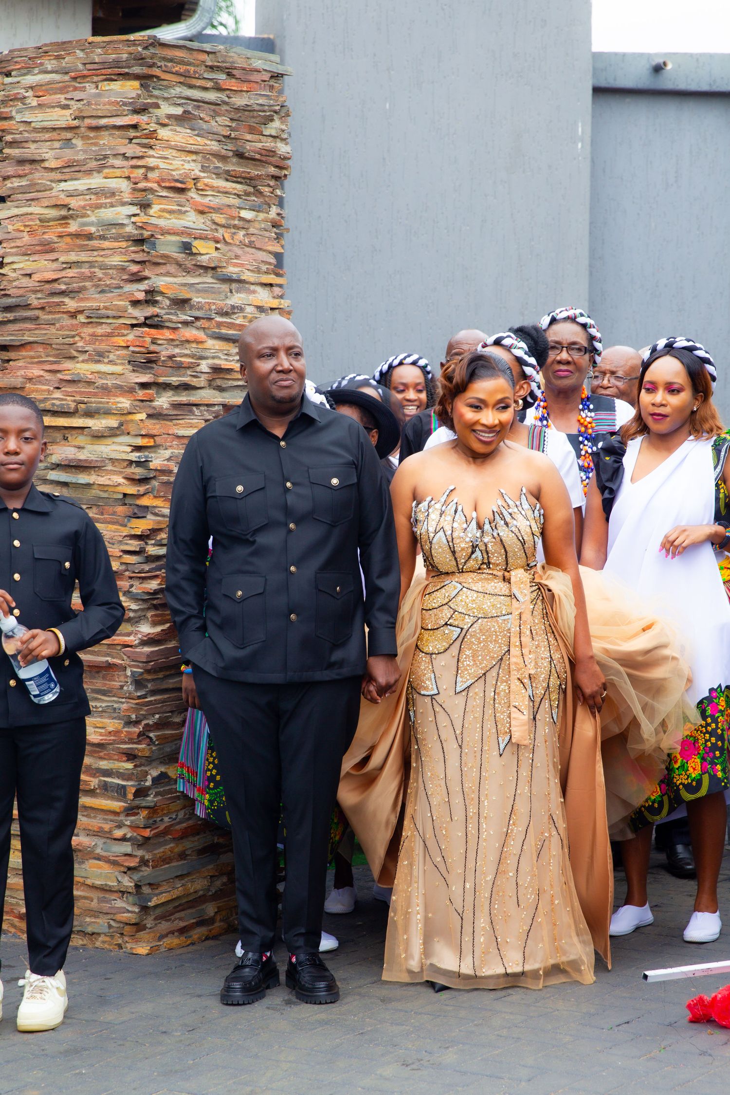 Bride in gold beaded gown and groom in black suit posing with wedding party at stone wall venue.