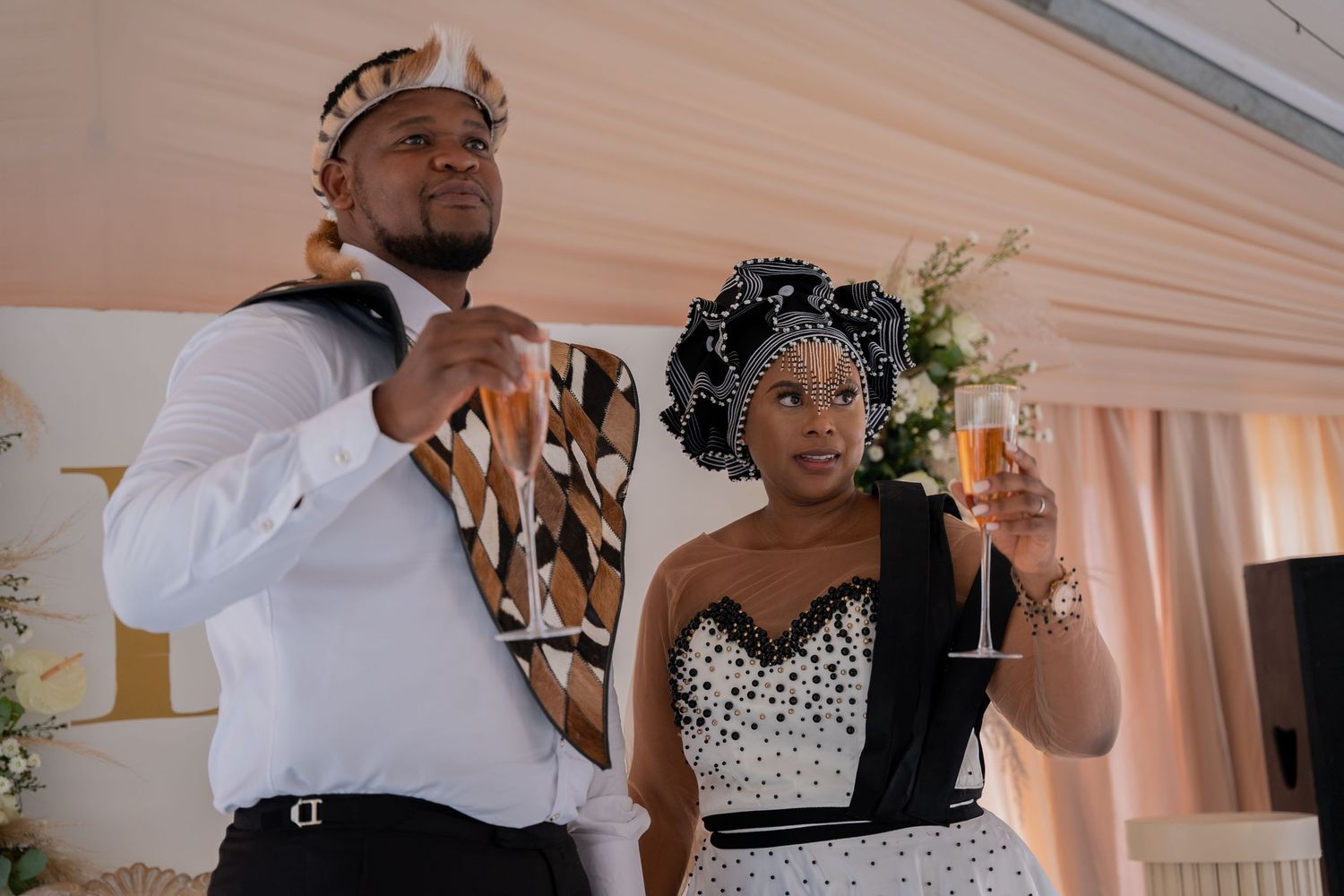 Couple in black and white traditional African attire raising champagne glasses at a wedding reception.